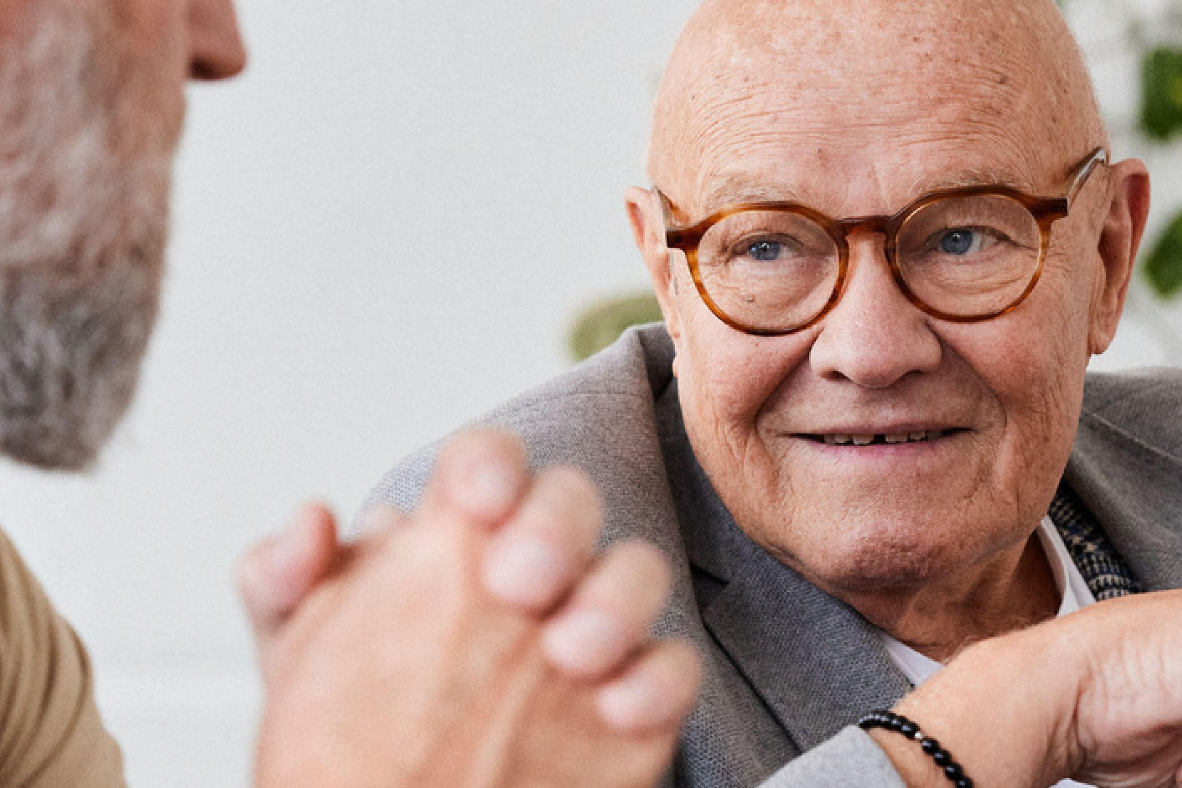 Close-up of an elderly man with round glasses and a warm smile, engaging in a conversation with another person whose hands and partial face are visible. The man is dressed in a blazer, exuding a sense of wisdom and friendliness in a cozy, light-filled environment. Ideal for showcasing themes of senior care, companionship, or meaningful conversations.
