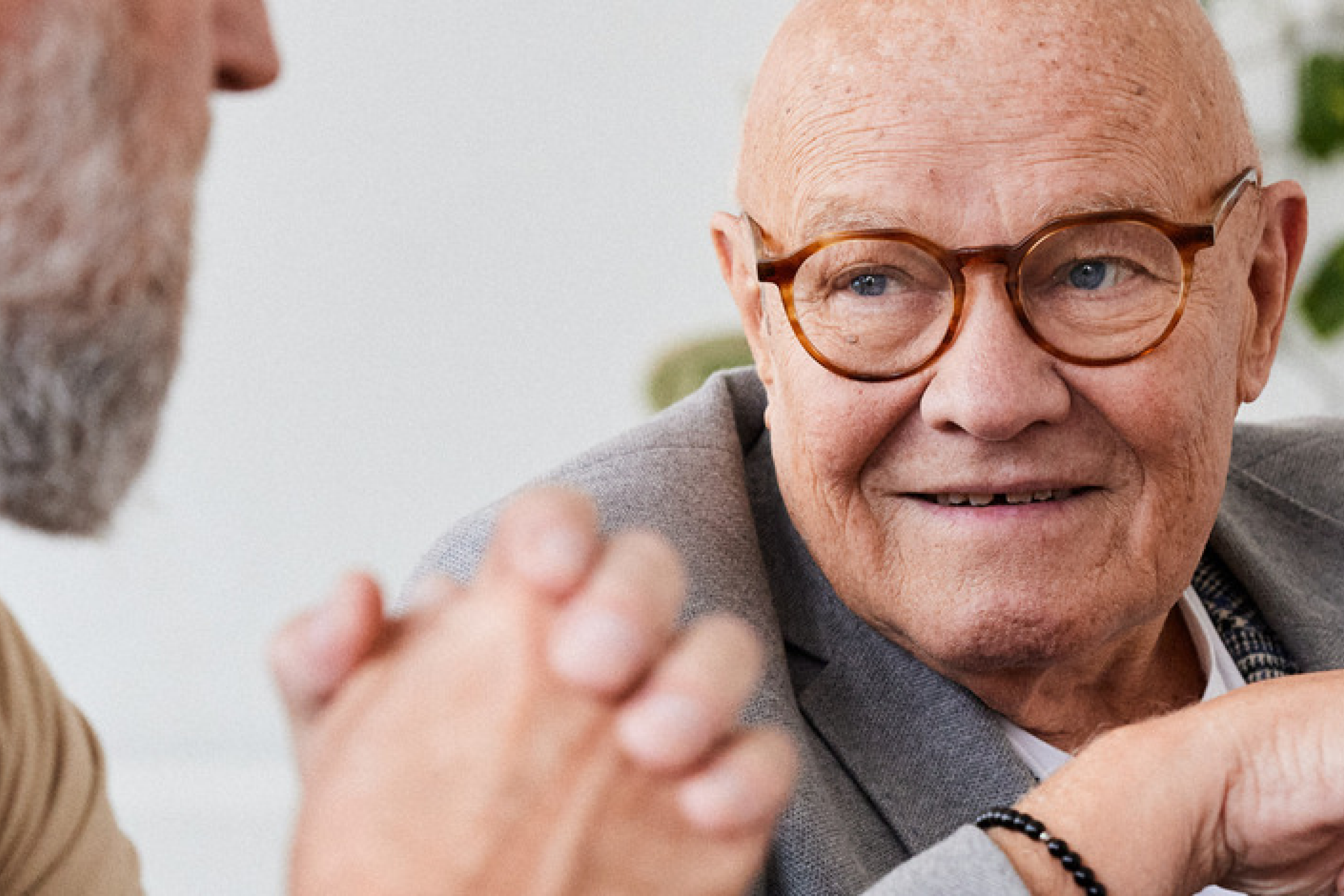 Close-up of an elderly man with round glasses and a warm smile, engaging in a conversation with another person whose hands and partial face are visible. The man is dressed in a blazer, exuding a sense of wisdom and friendliness in a cozy, light-filled environment. Ideal for showcasing themes of senior care, companionship, or meaningful conversations.