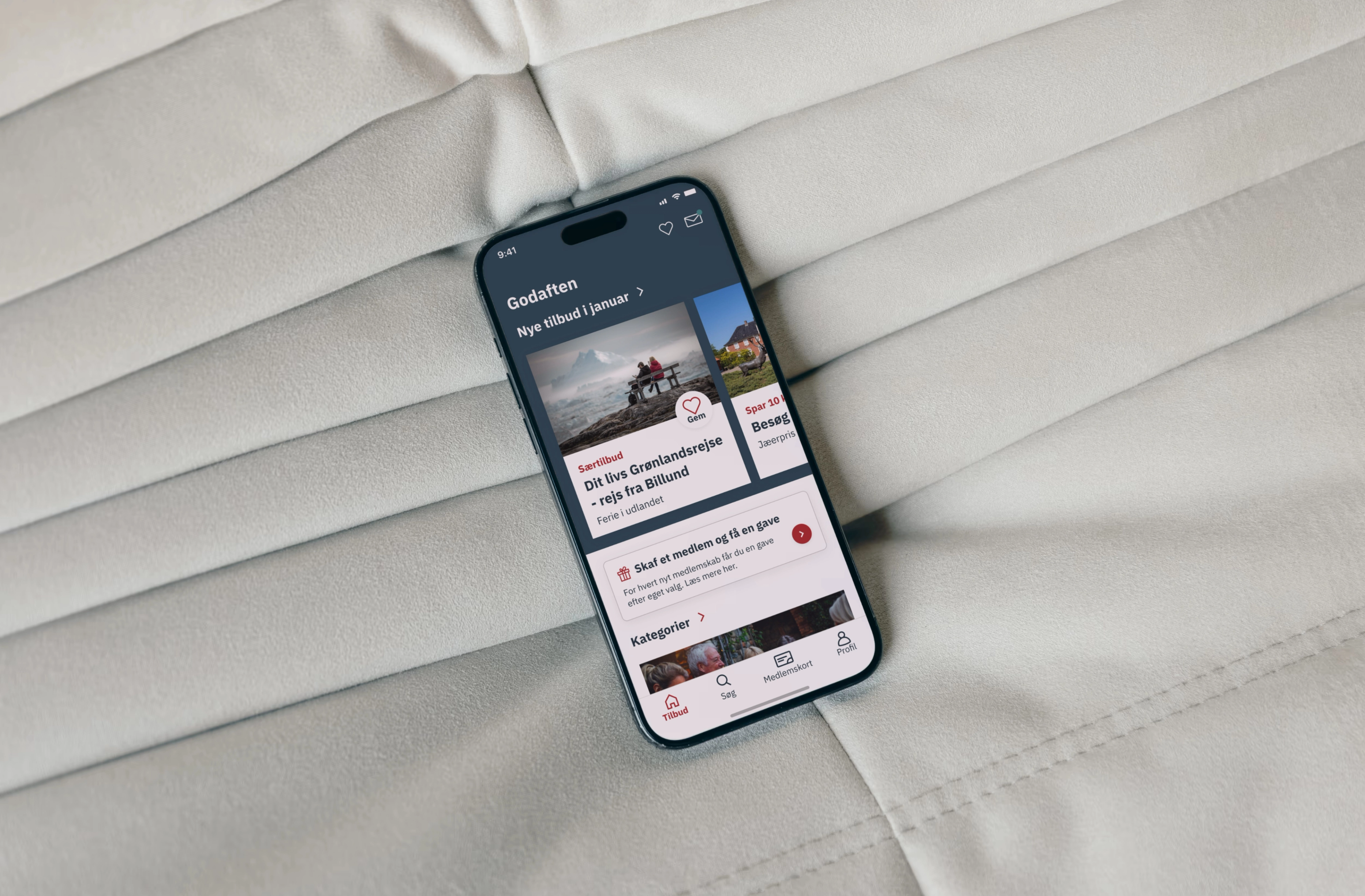 A smartphone displaying the Ældre Sagen app's landing page, resting on a soft gray comforter. The screen shows a clean and inviting app interface, with accessible buttons and a welcoming design tailored for all users.