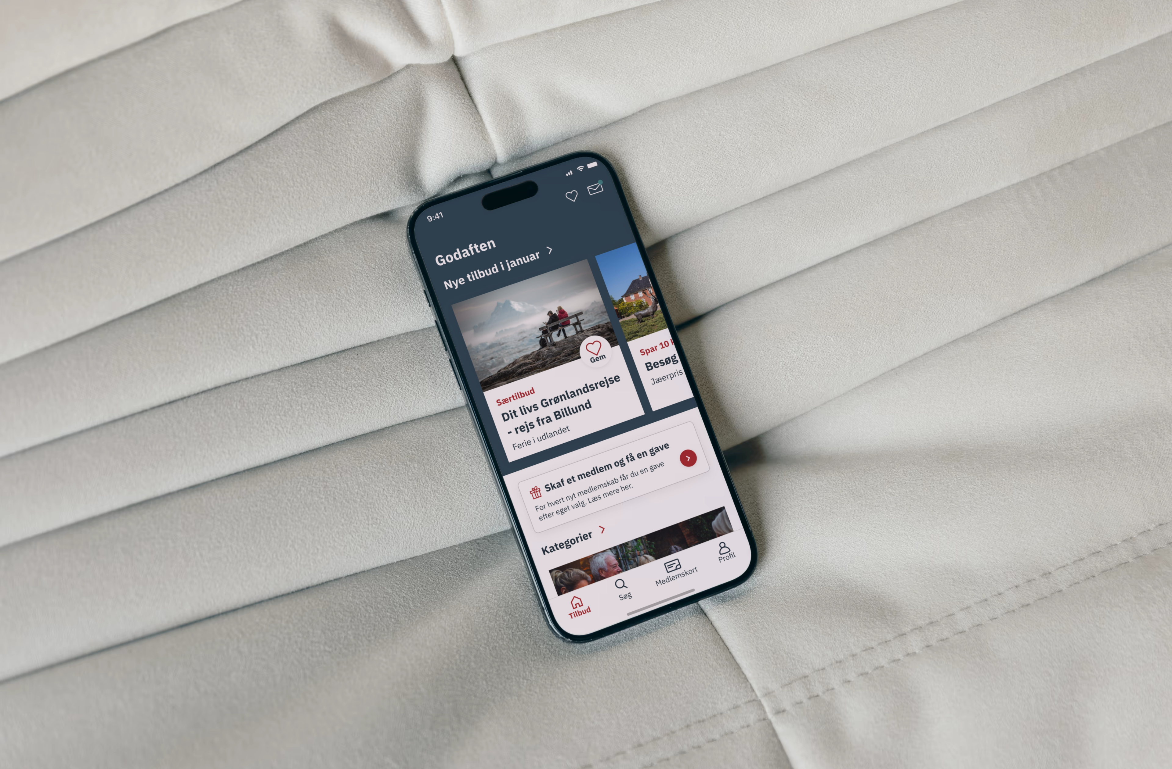 A smartphone displaying the Ældre Sagen app's landing page, resting on a soft gray comforter. The screen shows a clean and inviting app interface, with accessible buttons and a welcoming design tailored for all users.