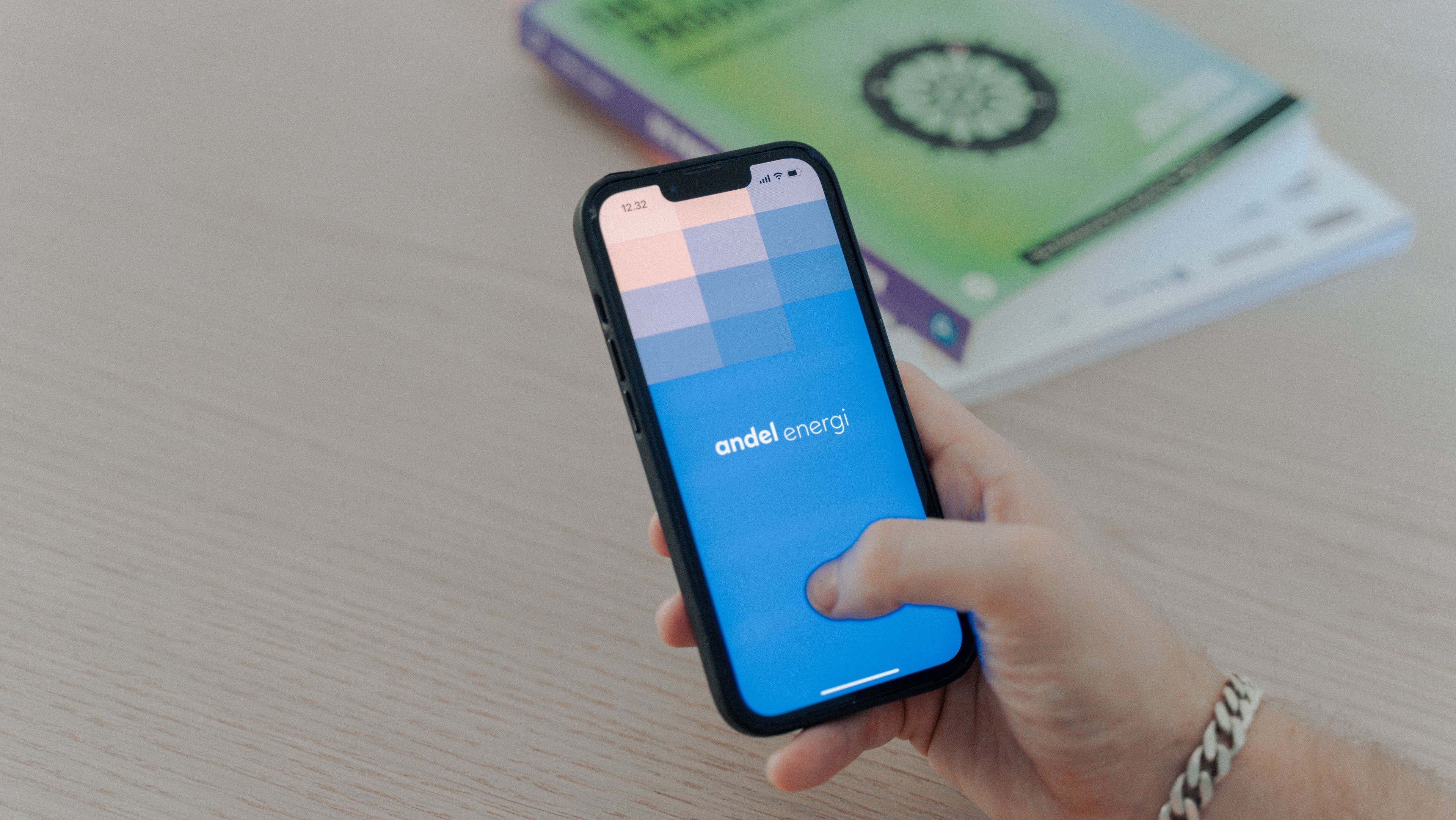 A person holding a smartphone displays the "andel energi" app on the screen, which features a blue gradient background with a checkered pattern at the top. The person is wearing a silver chain bracelet, and a green book is visible on the light wooden table in the background.