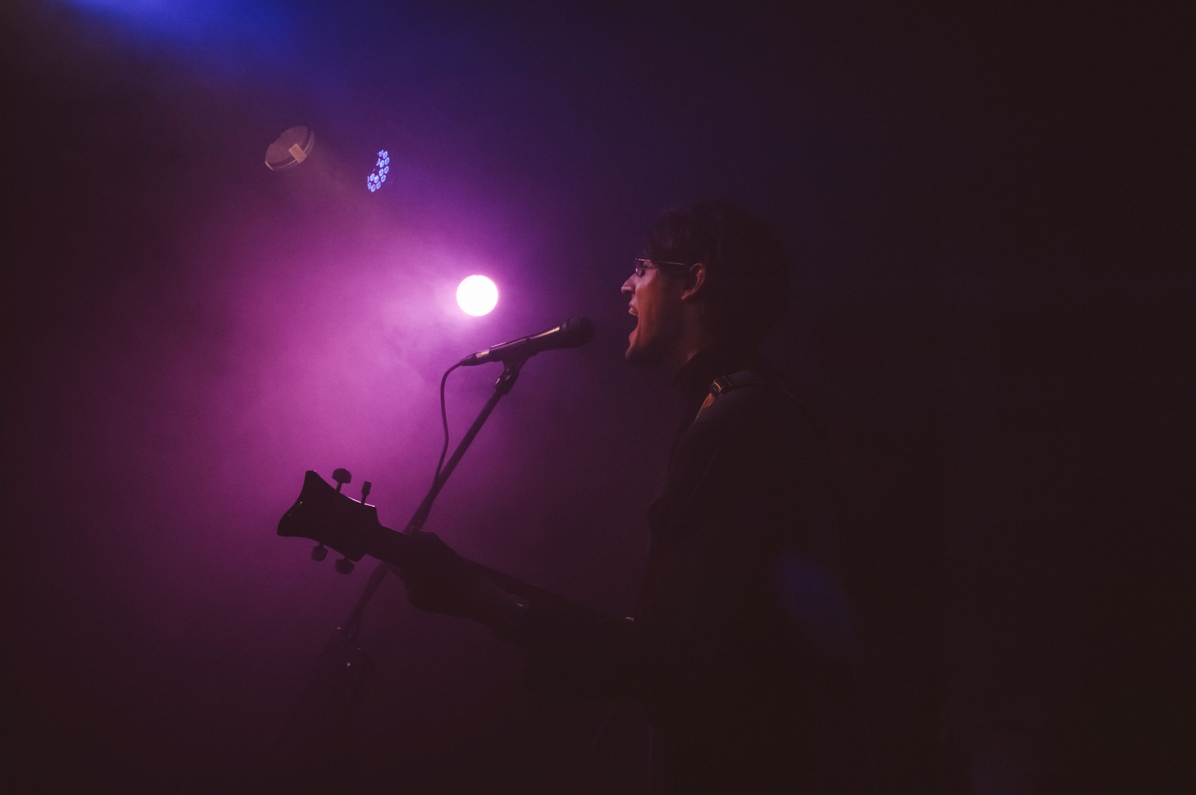 A musician passionately sings into a microphone while playing guitar on stage, illuminated by dramatic purple and blue stage lighting with a smoky atmosphere.