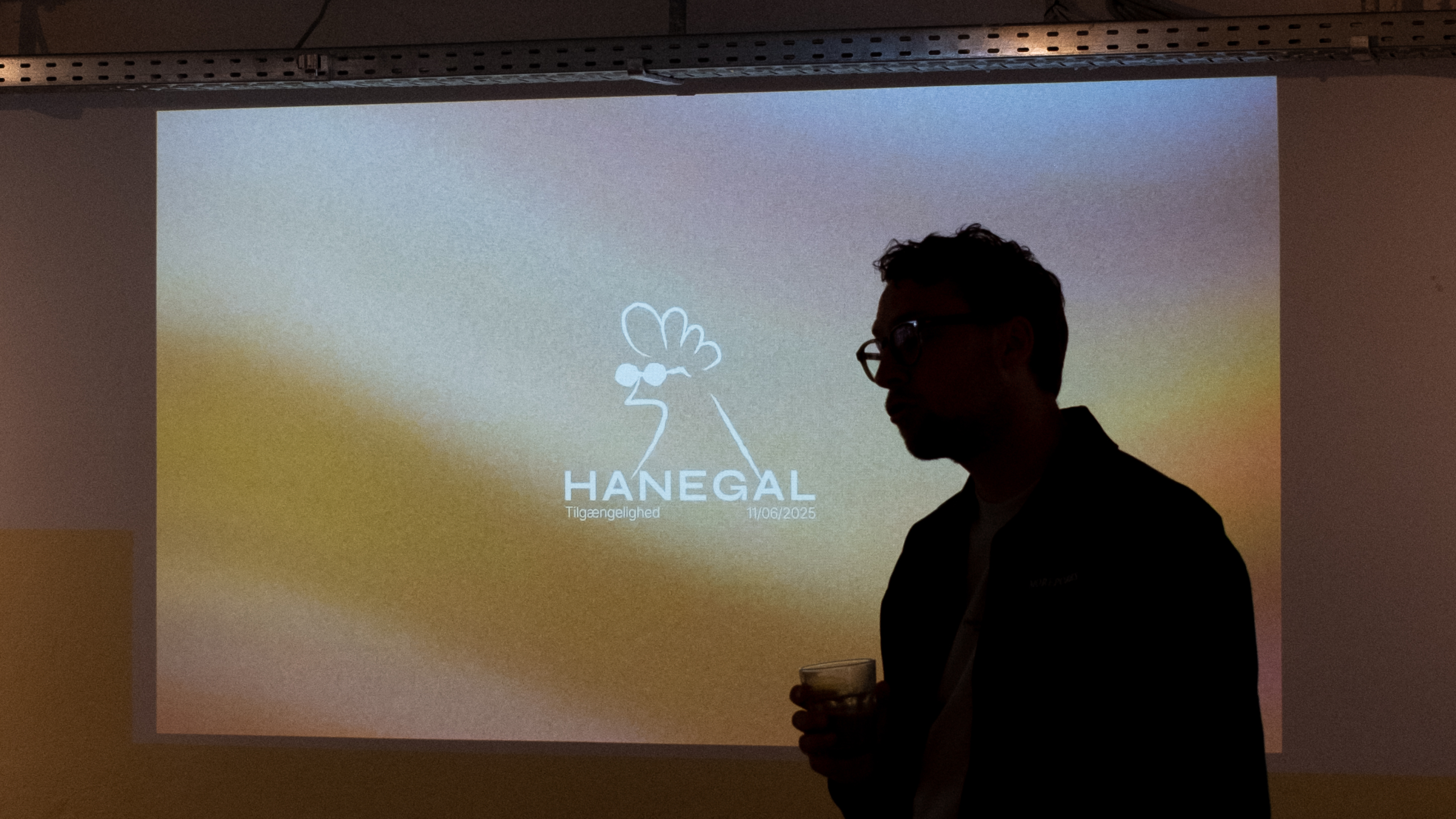 A silhouette of a person holding a drink in front of a projected screen displaying a logo with a stylized chicken wearing glasses, and the text "HANEGAL Tilgængelighed 11/06/2025." The room is dimly lit, and the focus is on the projection.