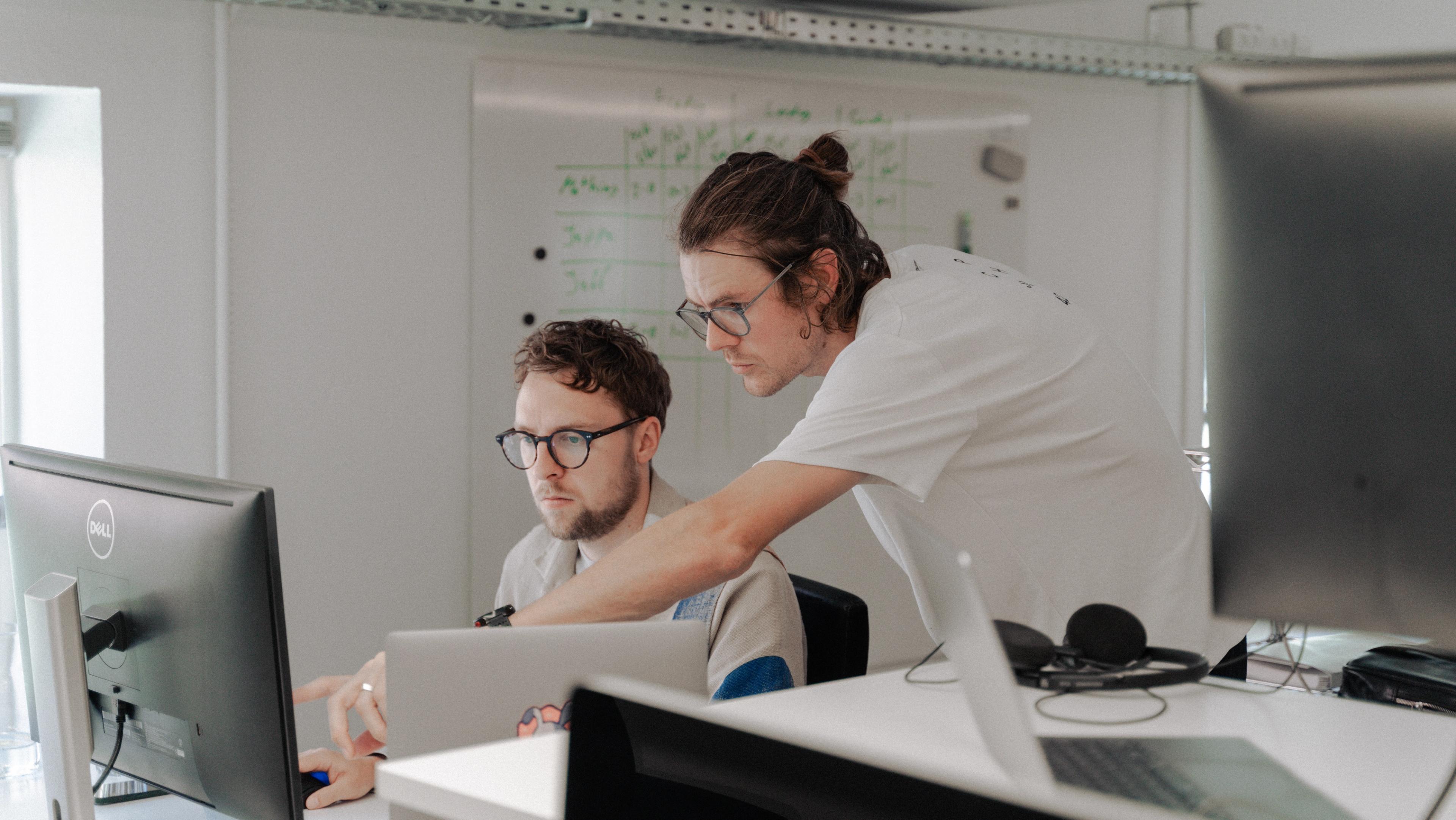 Two men looking at a computer by a work desk. One of them point at the screen as a way of explaining