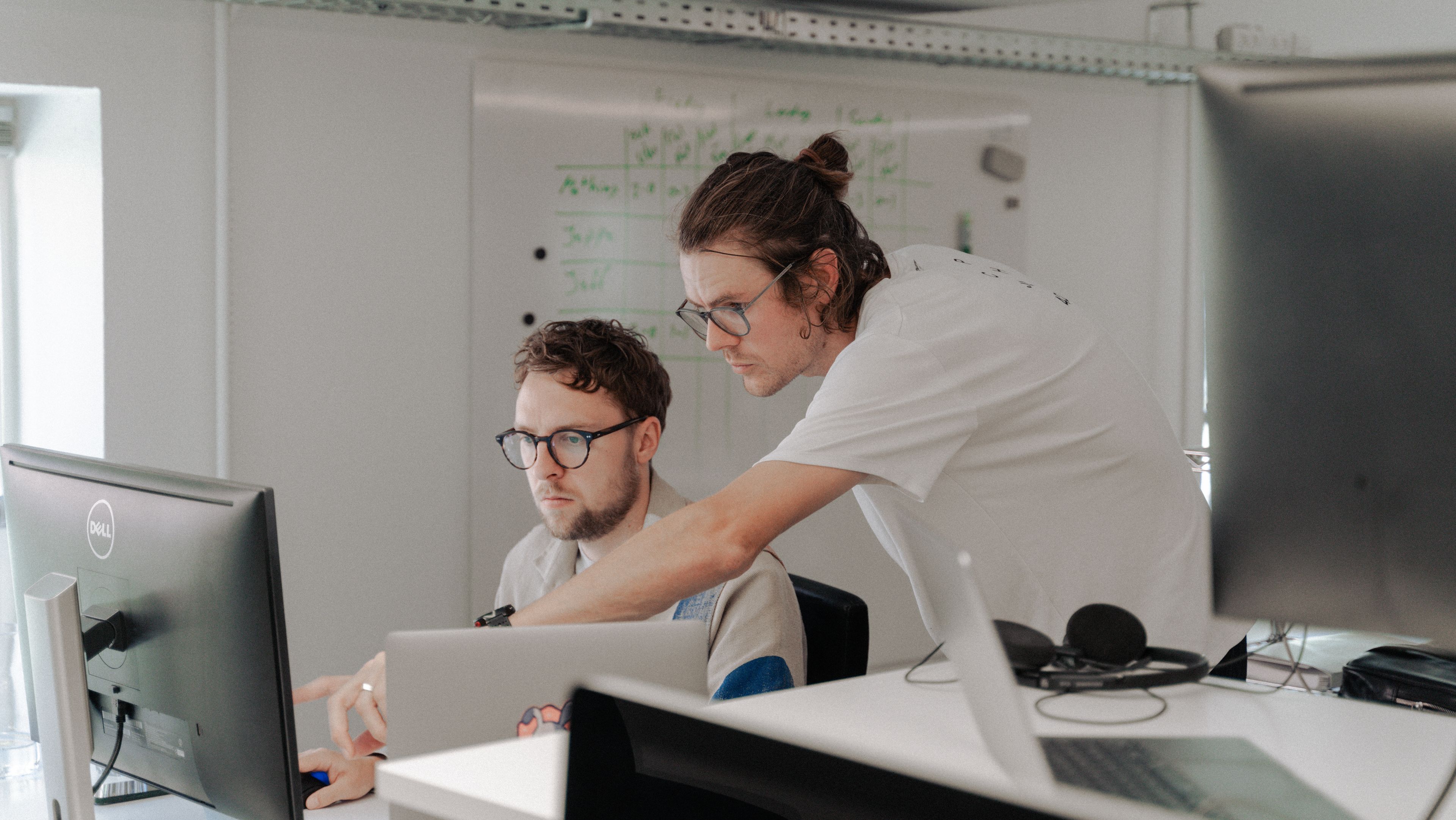 Two men looking at a computer by a work desk. One of them point at the screen as a way of explaining