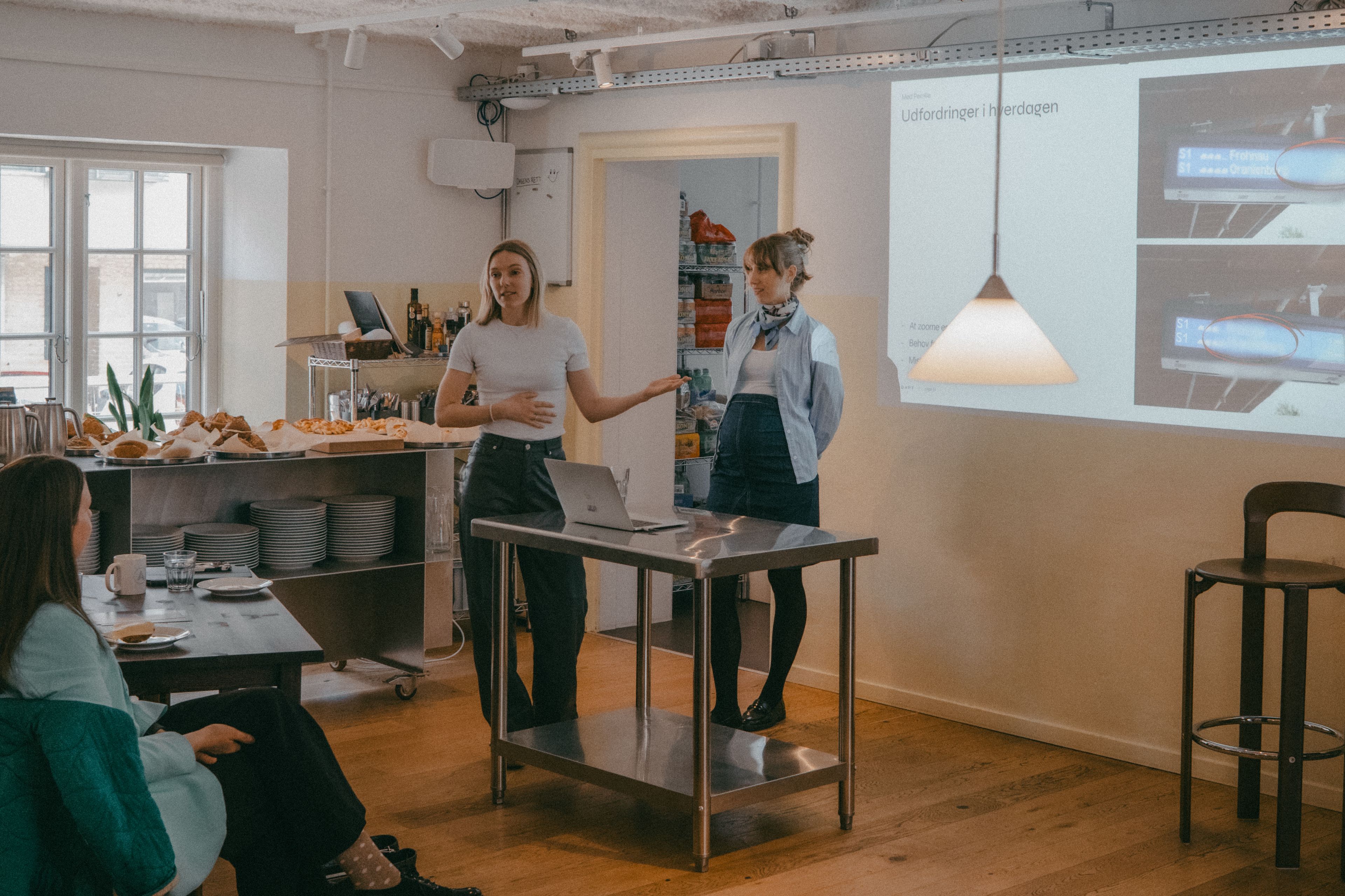 Two women are giving a presentation in a bright room with a kitchen setting. One woman is gesturing while speaking, standing beside a laptop on a stainless steel table. Several people are seated and attentively listening.