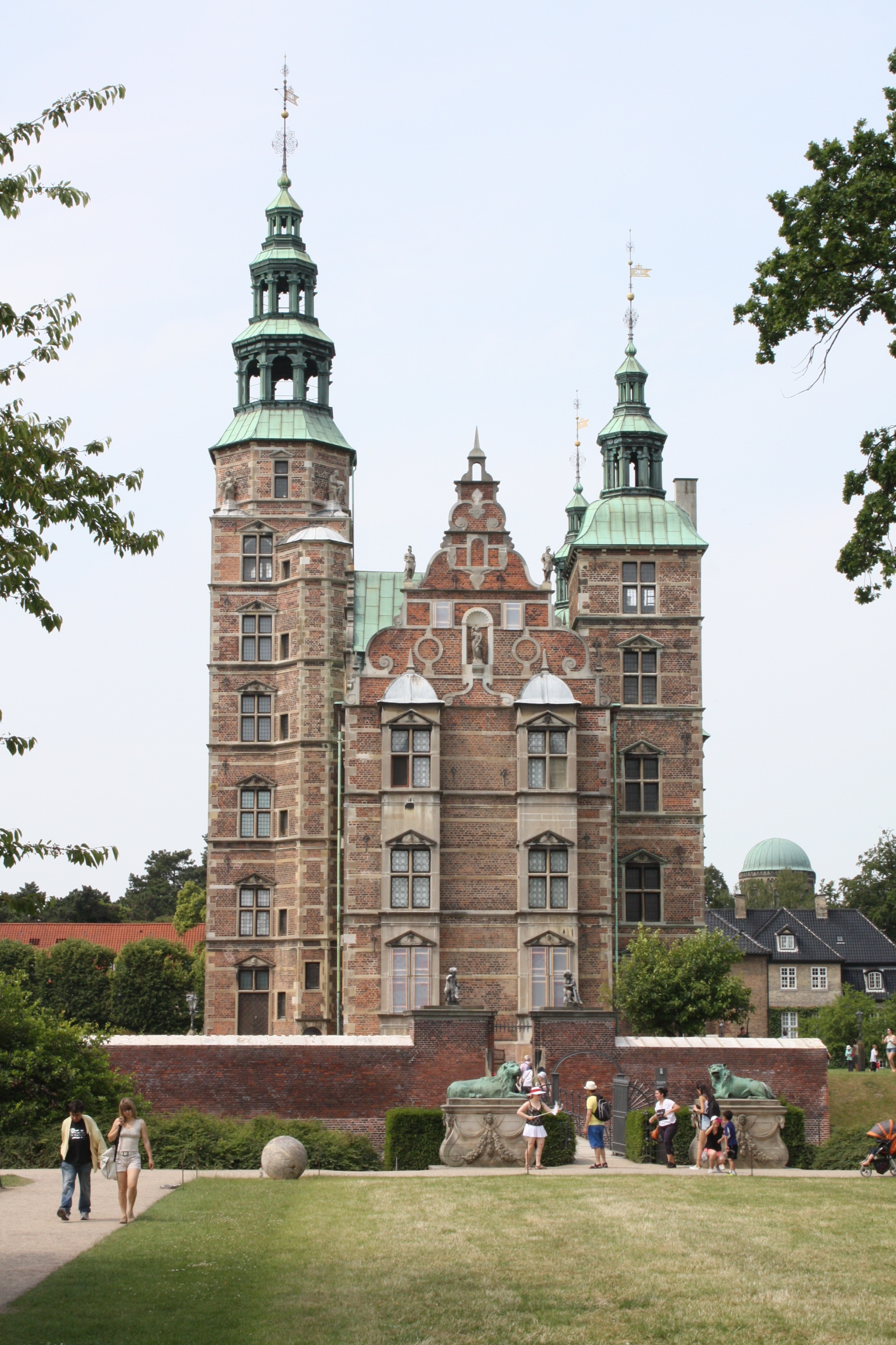 Rosenborg Slot in Copenhagen stands majestically under a bright blue sky, surrounded by lush green trees with vibrant leaves. The well-manicured green grass in the foreground adds to the picturesque scene, while a few tourists can be seen admiring the historic castle, capturing its timeless charm.