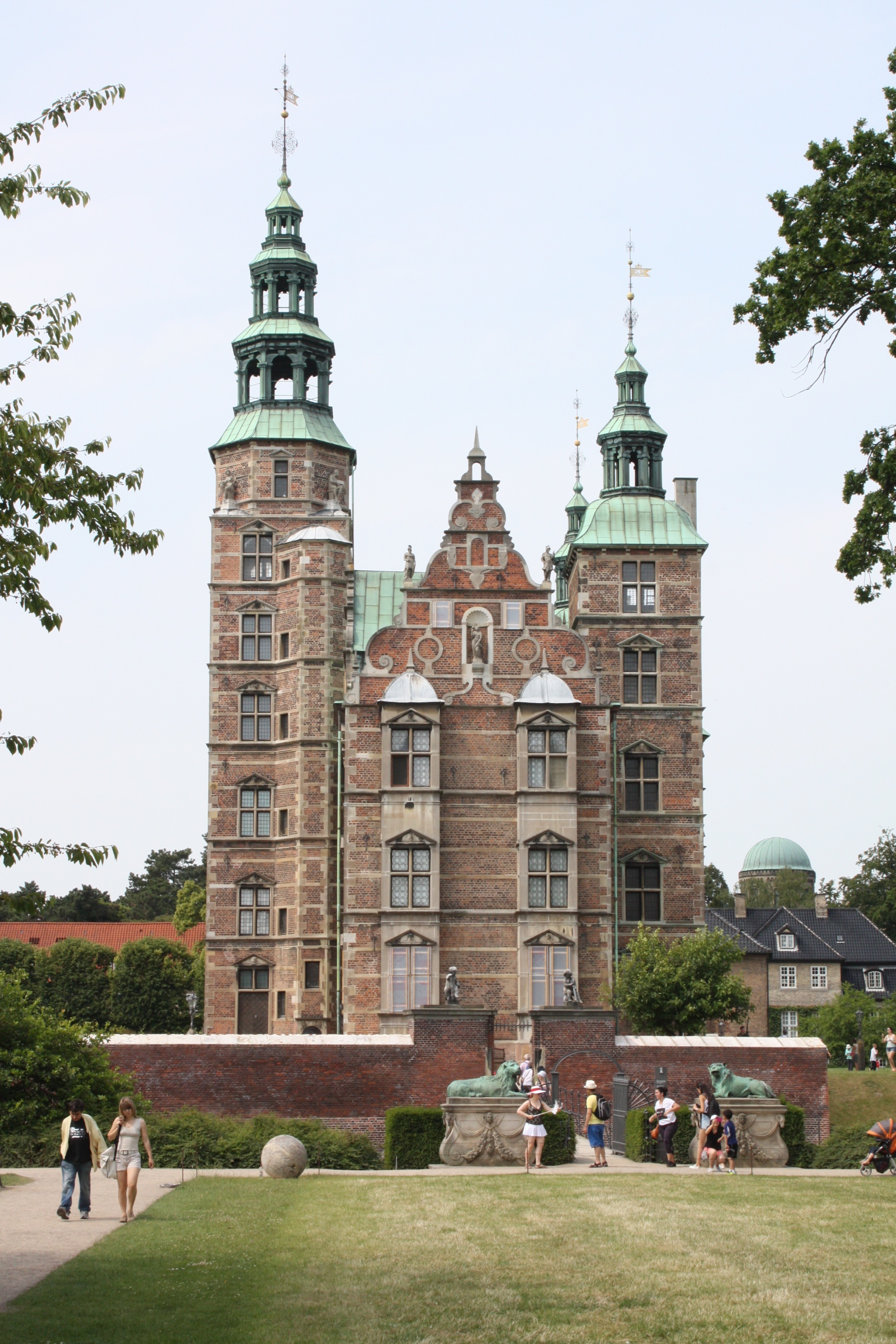 Rosenborg Slot in Copenhagen stands majestically under a bright blue sky, surrounded by lush green trees with vibrant leaves. The well-manicured green grass in the foreground adds to the picturesque scene, while a few tourists can be seen admiring the historic castle, capturing its timeless charm.