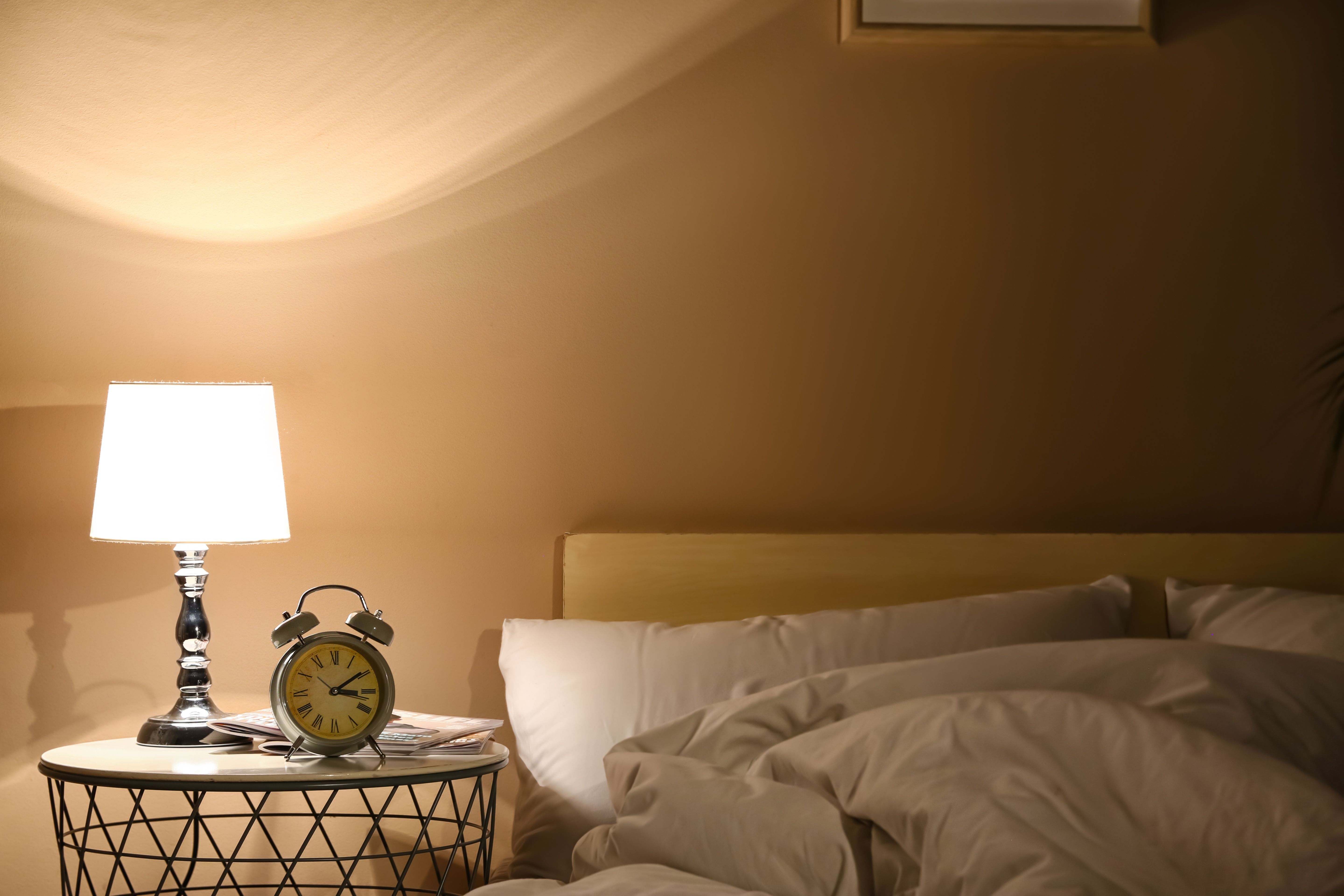 A lit bedside lamp and an alarm clock on a table beside an empty, turned down bed at night.