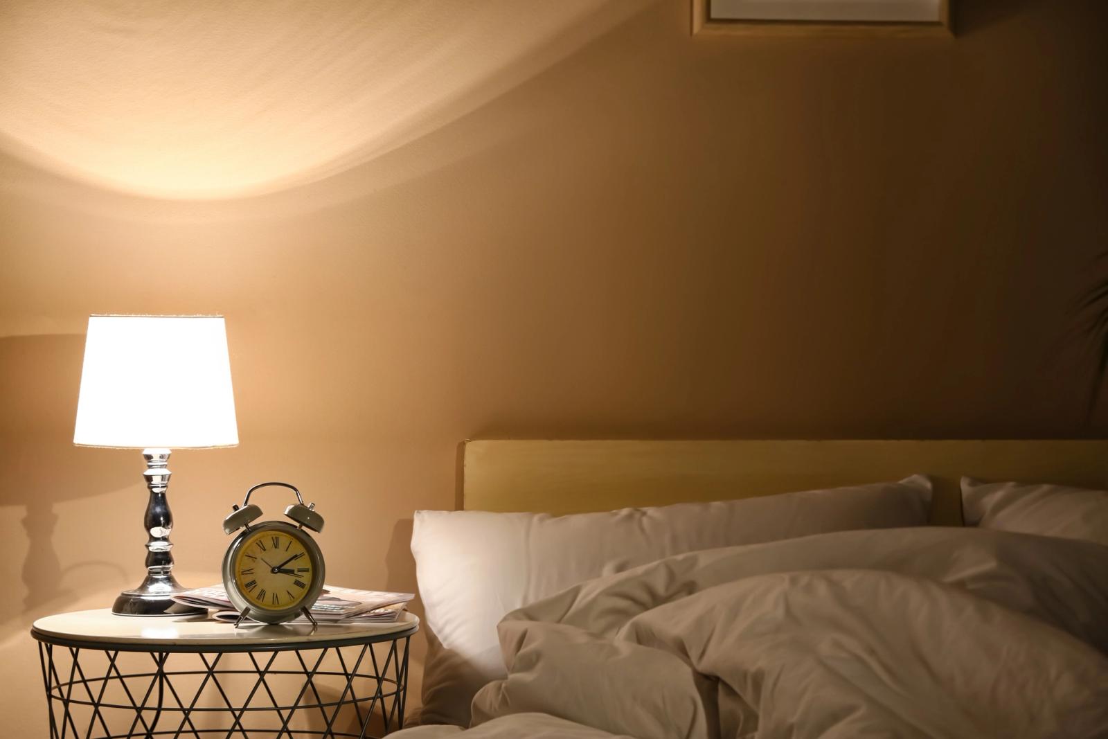 A lit bedside lamp and an alarm clock on a table beside an empty, turned down bed at night.