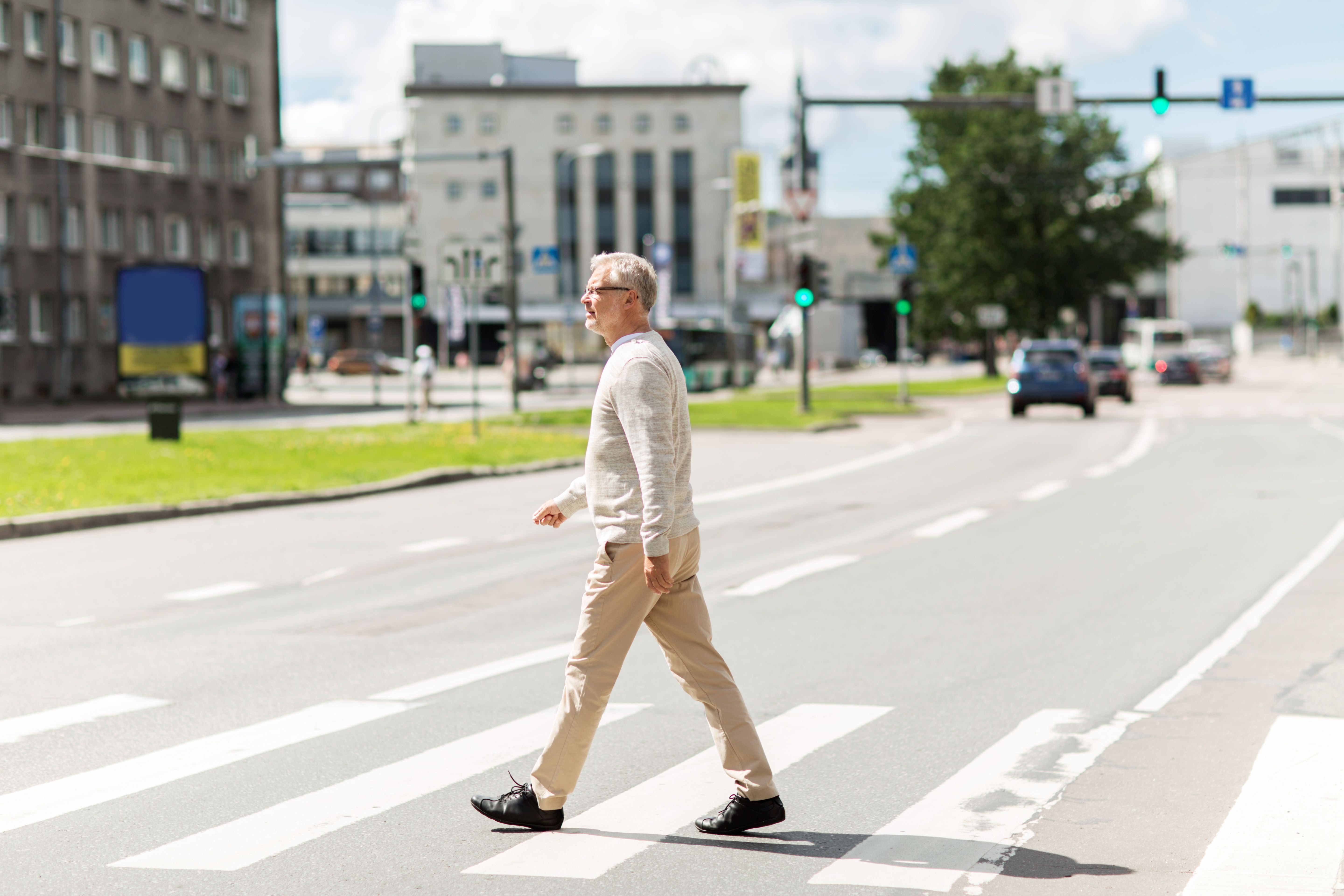 An older man in a pale shirt walking across a marked city crosswalk on a summer day, traffic and office buildings behind him.