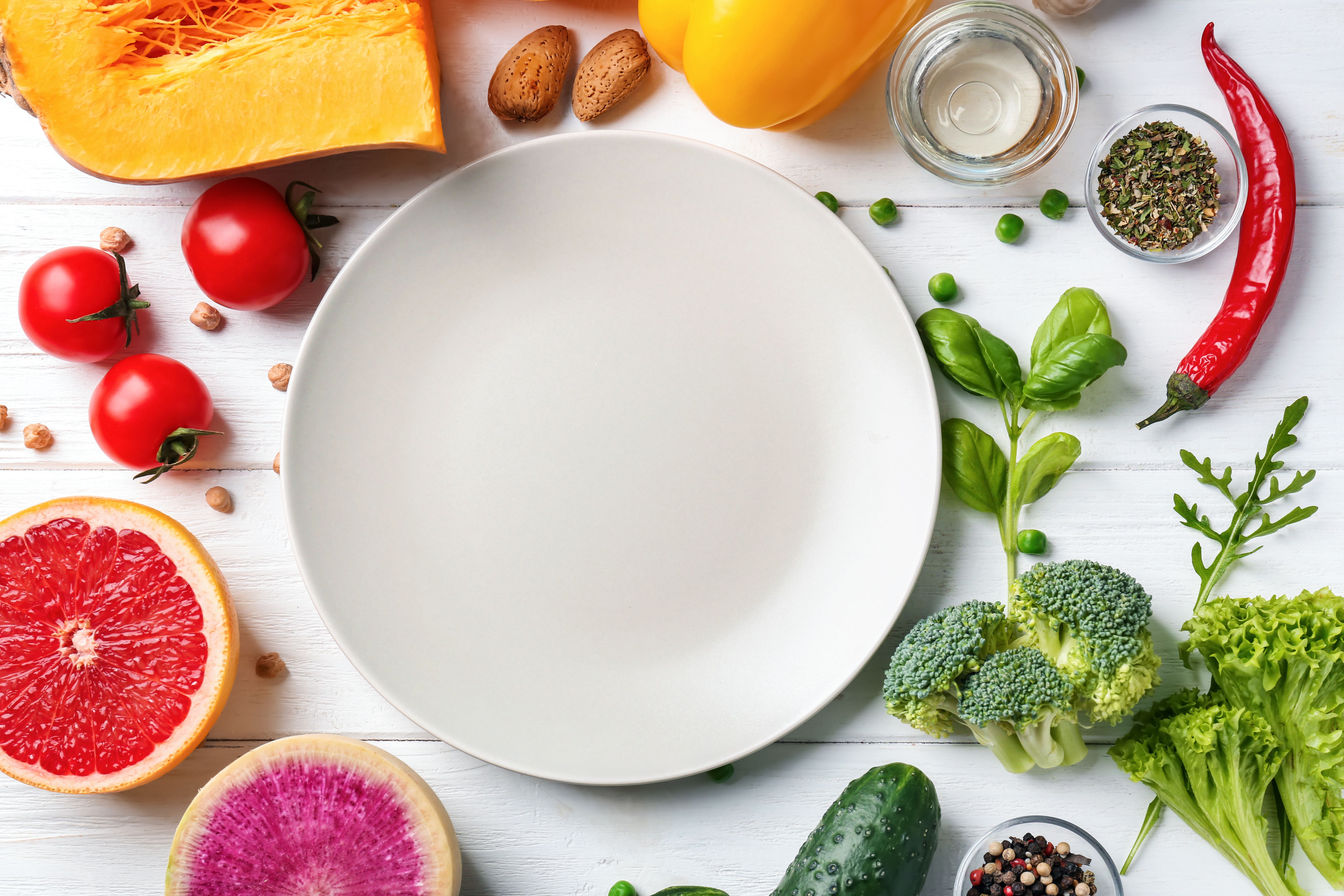 Fresh vegetables and fruit arranged around an empty white plate on a pale wooden table.
