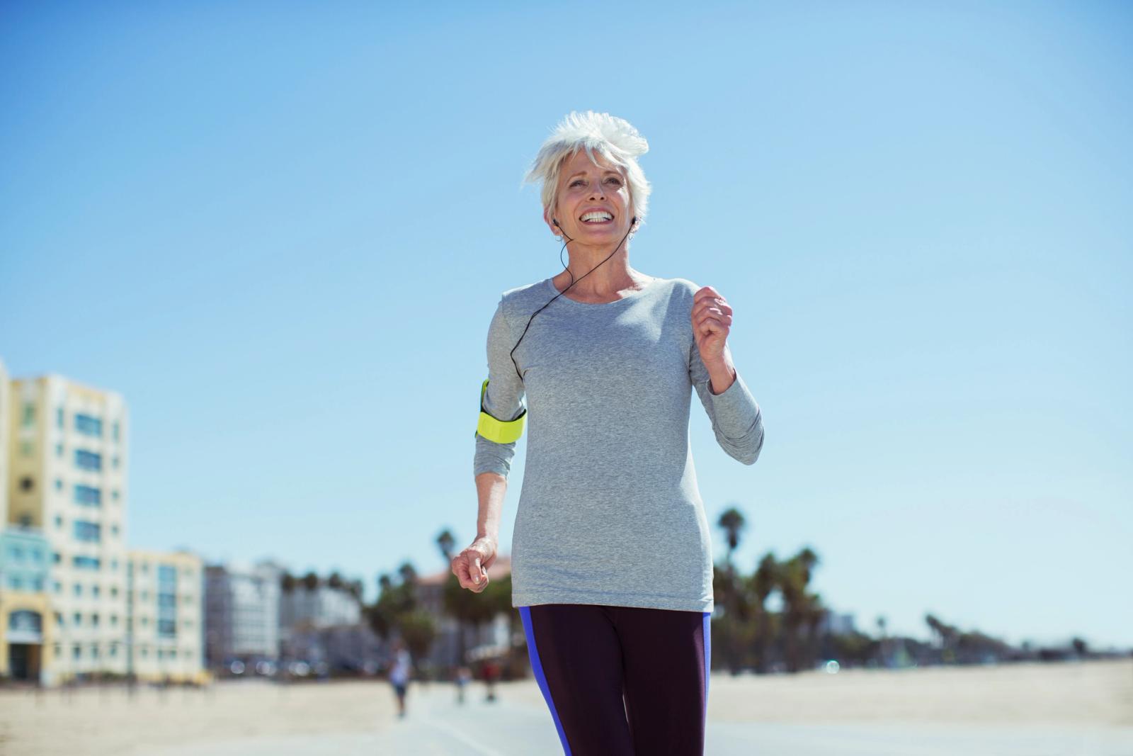 A grey-haired woman in a light top and dark leggings walking briskly along a sunlit beach promenade, arms swinging, with the sea and low buildings behind her.