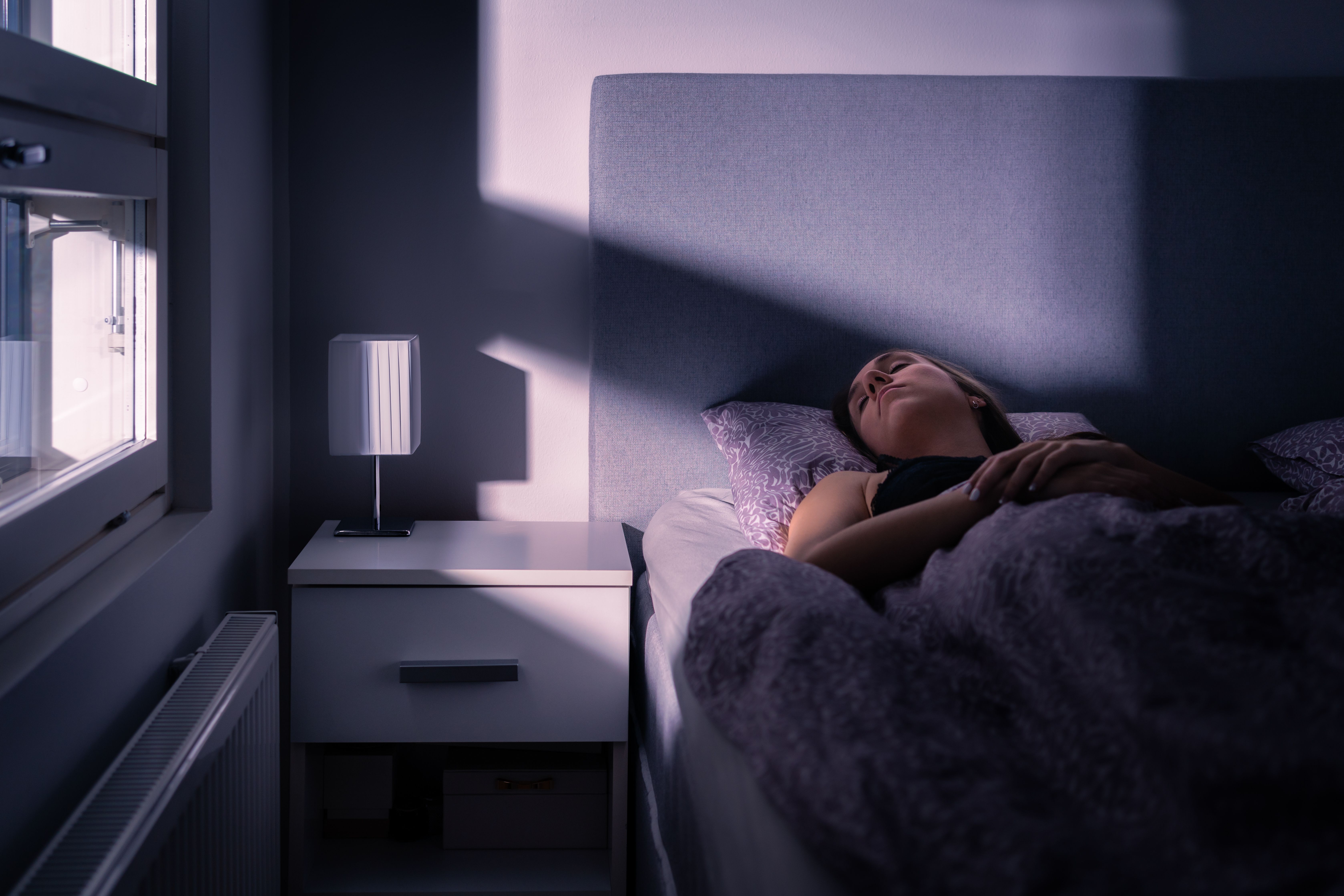 A woman asleep in bed in a dark bedroom, with a band of light from the window falling across the wall, bedside table and covers.