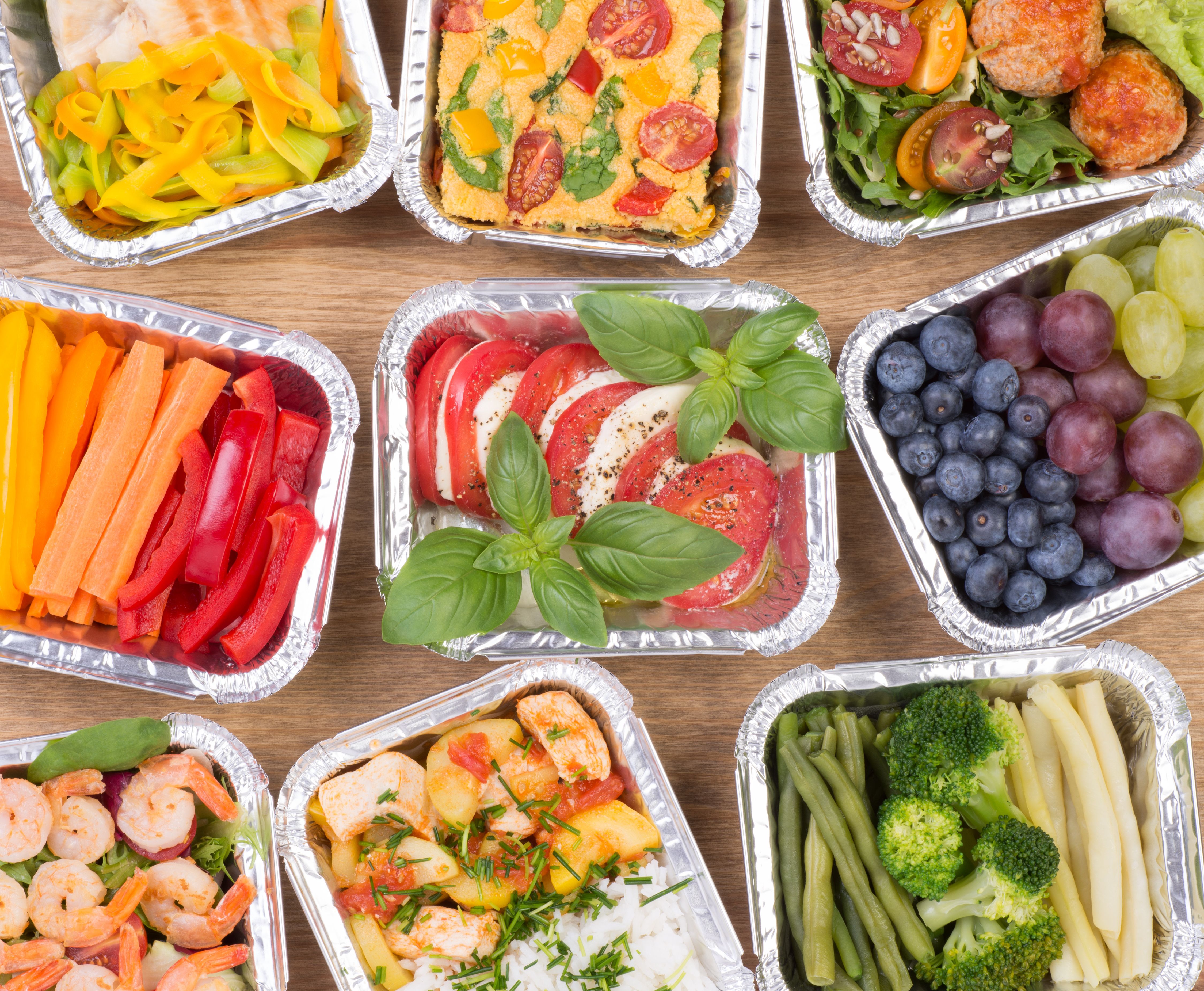 An assortment of prepared meals in foil takeaway containers seen from above on a wooden table: vegetables, salads, grains, fruit and cooked dishes.