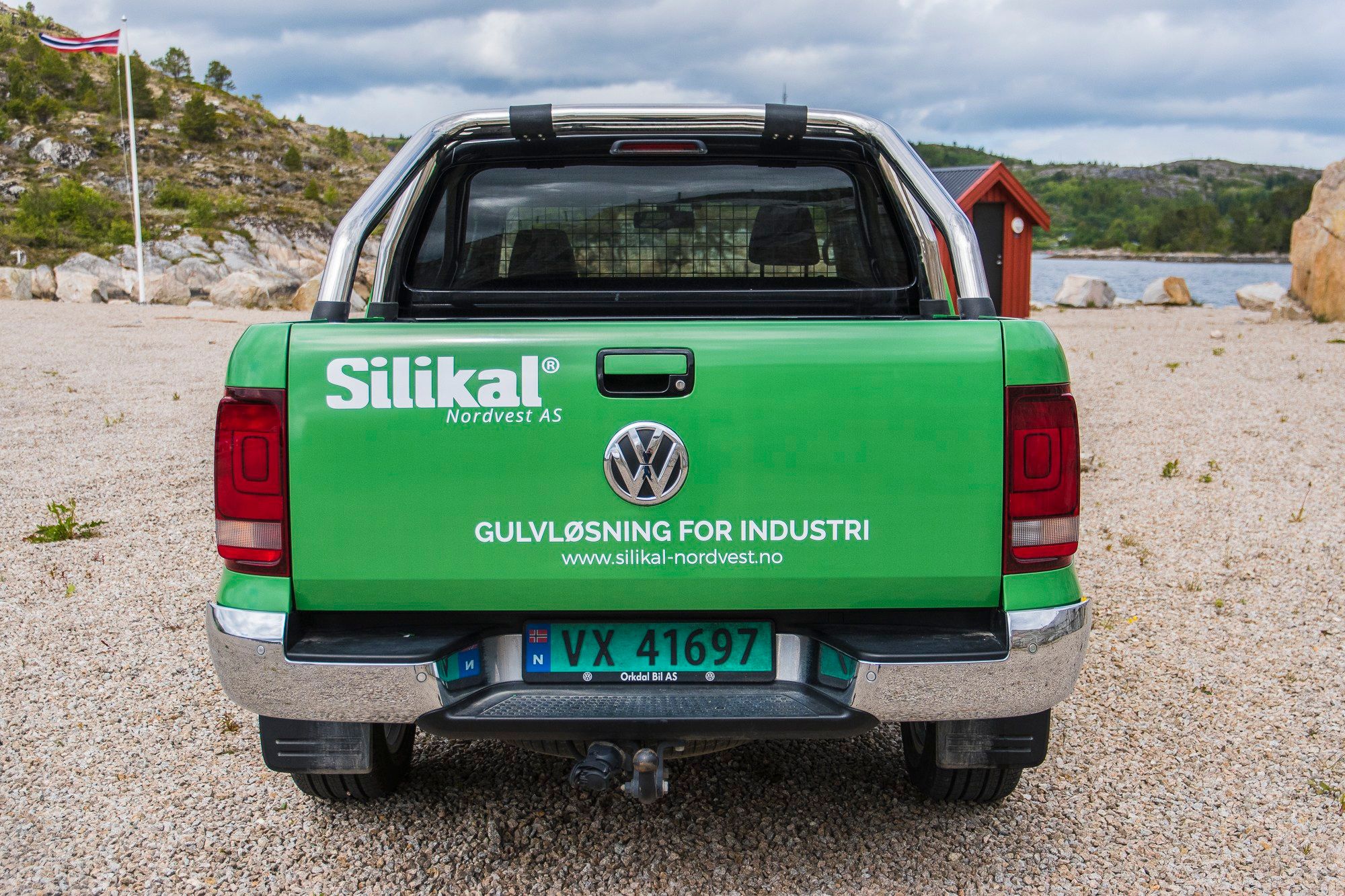 Grønn Volkswagen pick-up med Silikal Nordvest AS logo og teksten "Gulvløsning for industri" på lasteplanet, parkert ved strand.