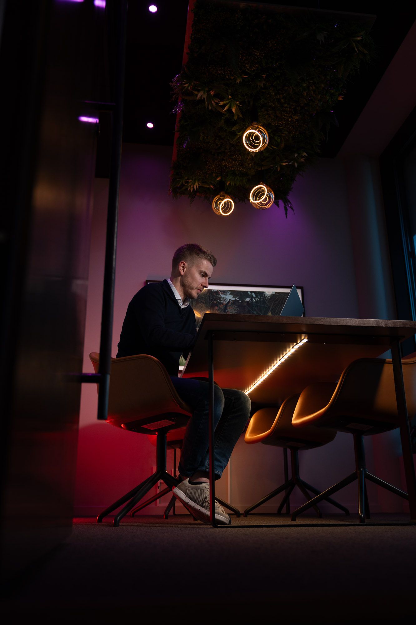 Mann ved skrivebord med neonbelysning arbeider på bærbar PC i moderne kontorrom med lilla og orange lysambiance.