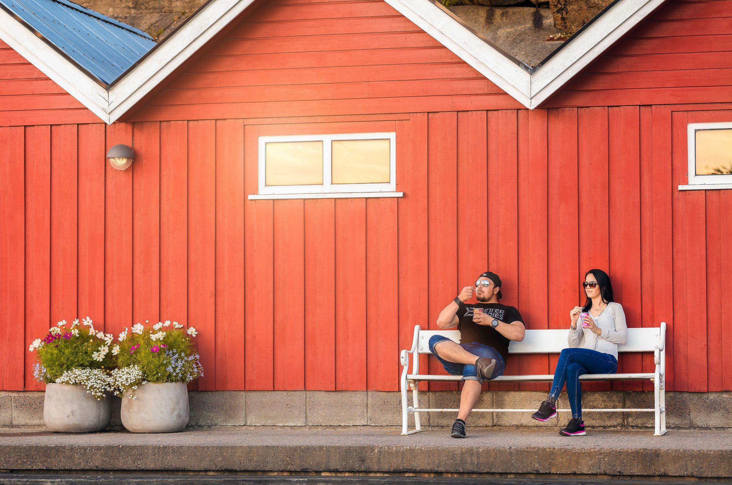 To personer sitter på en hvit benk foran en rødt hus med hvite vinduer og blomsterkrukker på fortauet.