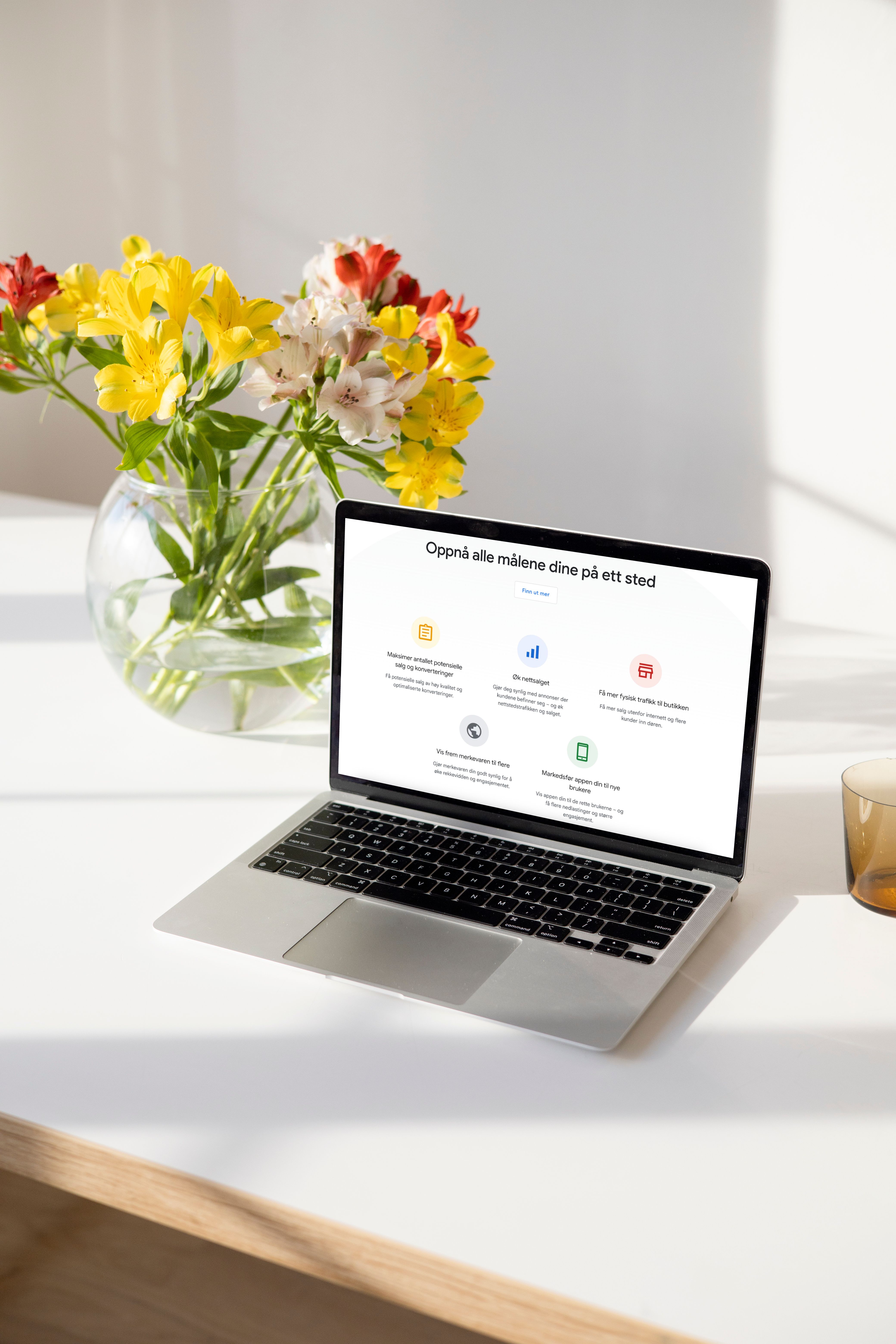 Laptop viser nettside med overskriften "Oppnå alle målene dine på ett sted" ved siden av blomster i vase og kaffe.