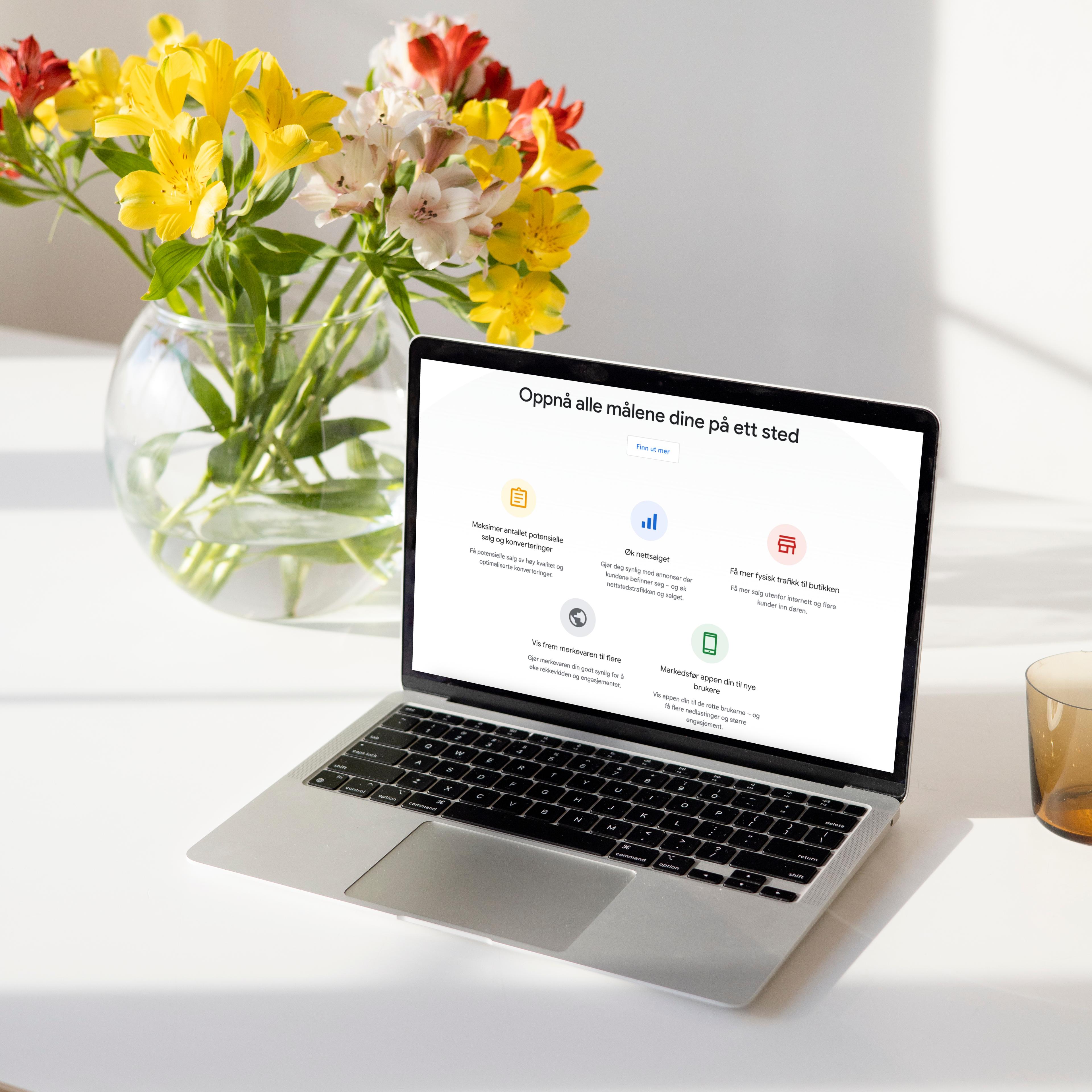 Laptop viser nettside med overskriften "Oppnå alle målene dine på ett sted" ved siden av blomster i vase og kaffe.