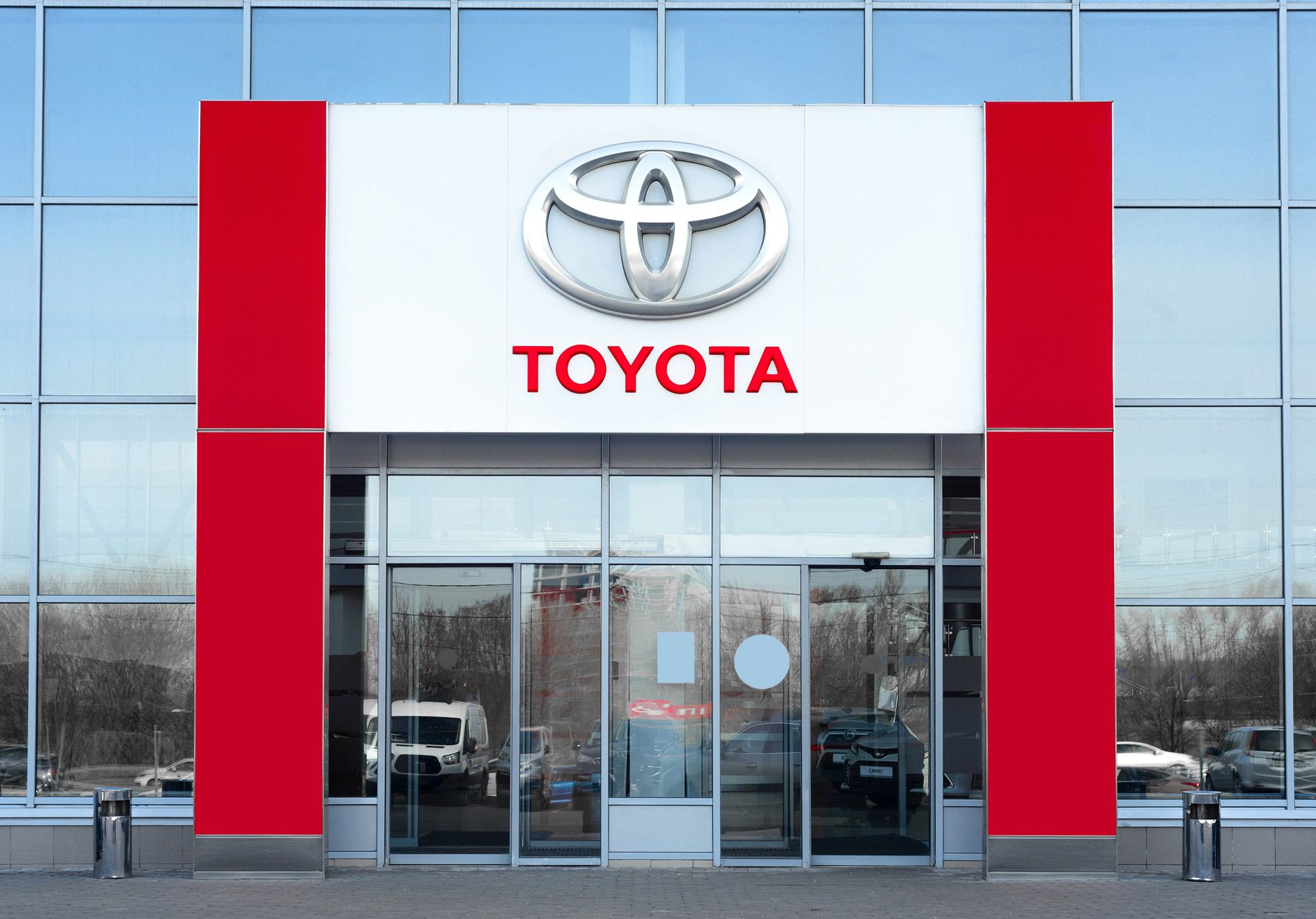 Toyota-forhandler med rødt og hvitt skilt, logo og navn på moderne bygning med glassfront og parkerte biler.