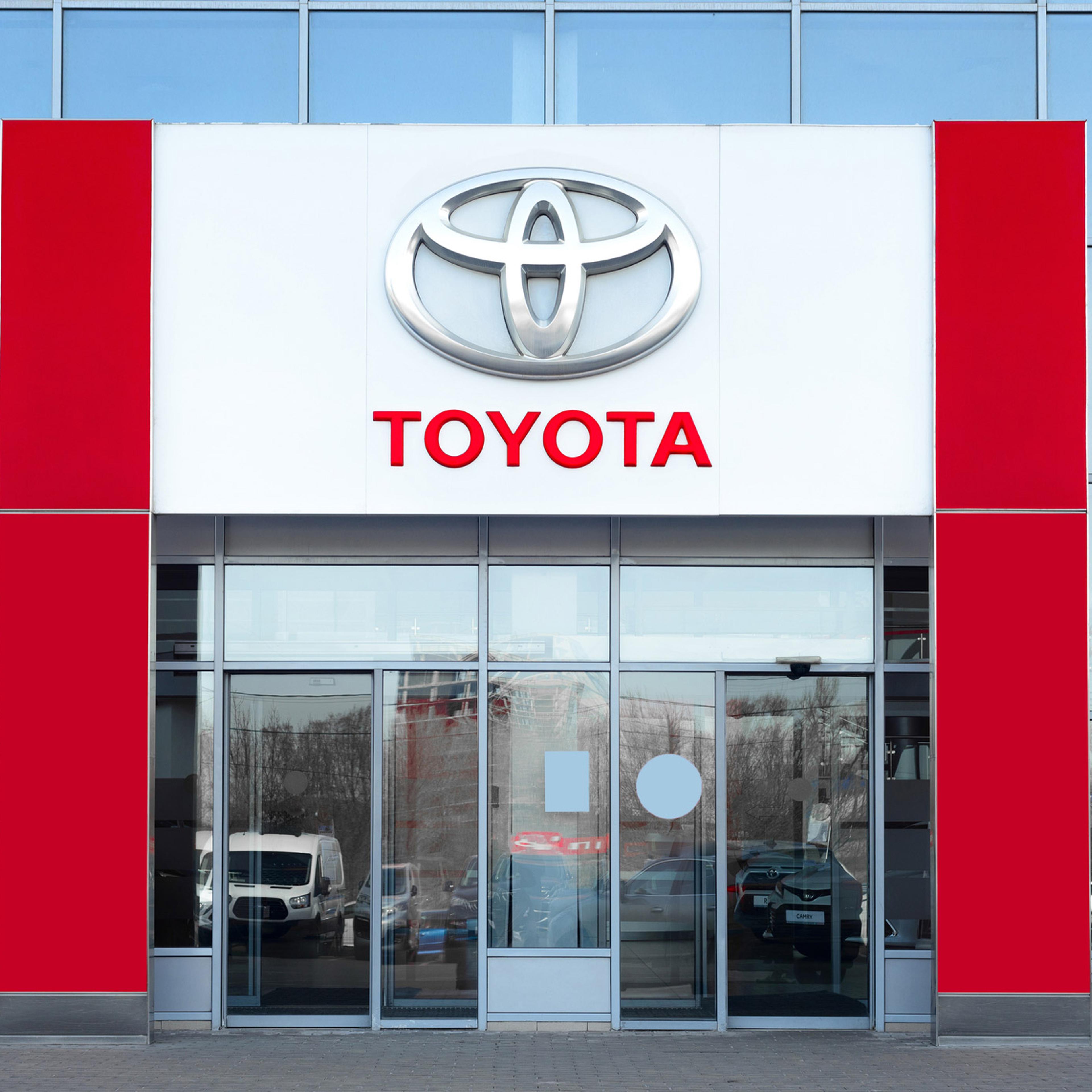 Toyota-forhandler med rødt og hvitt skilt, logo og navn på moderne bygning med glassfront og parkerte biler.