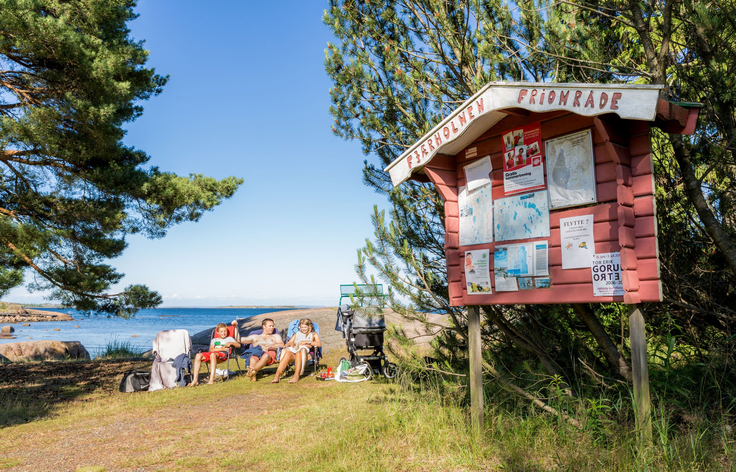 Familie ved strand med informasjonstavle merket "Priomsfjorde" i forgrunnen, blå hav og skog i bakgrunnen.