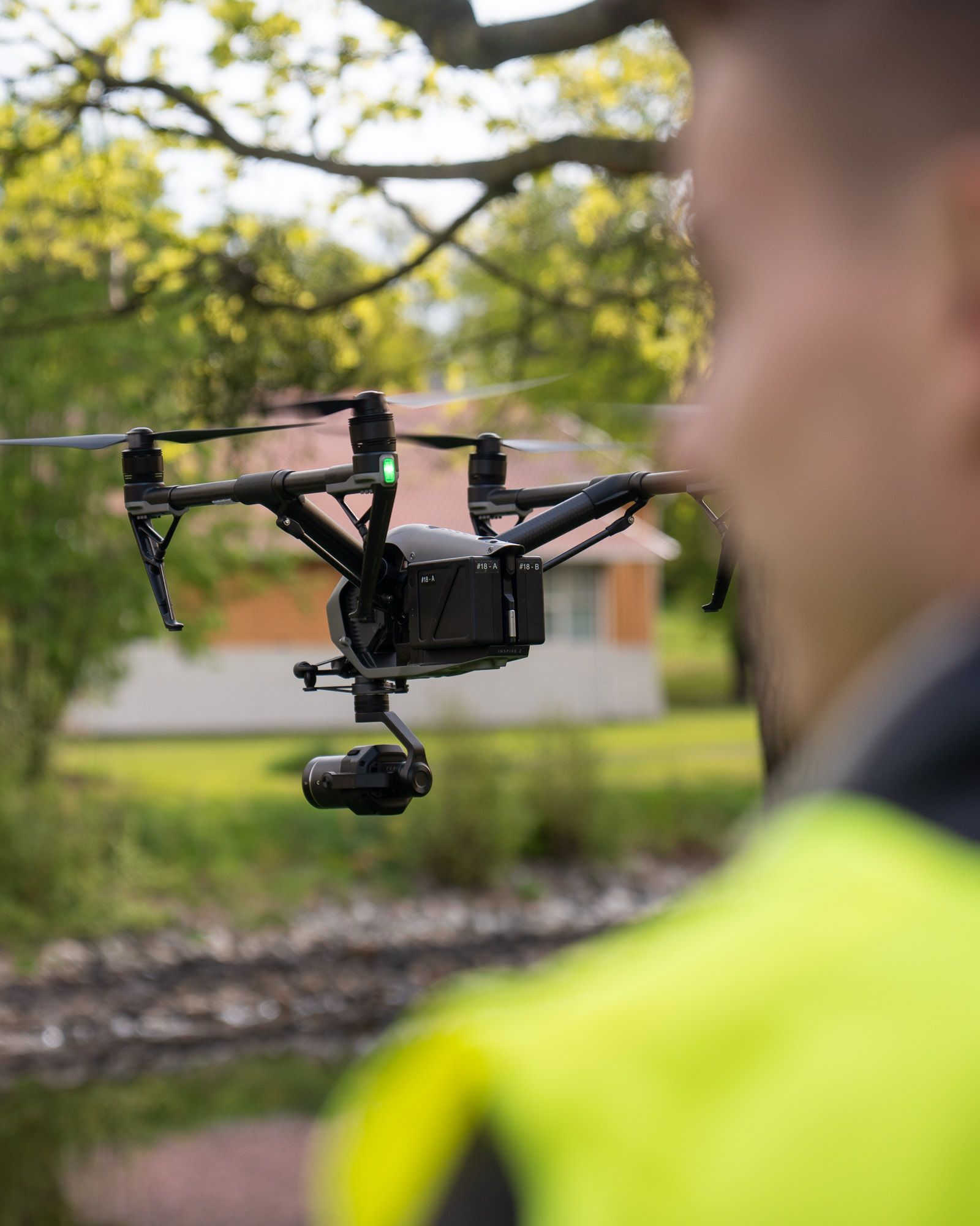 Drone-operatør i gul refleksvest styrer en svart multikopter over grønt landskap med bygning i bakgrunnen.