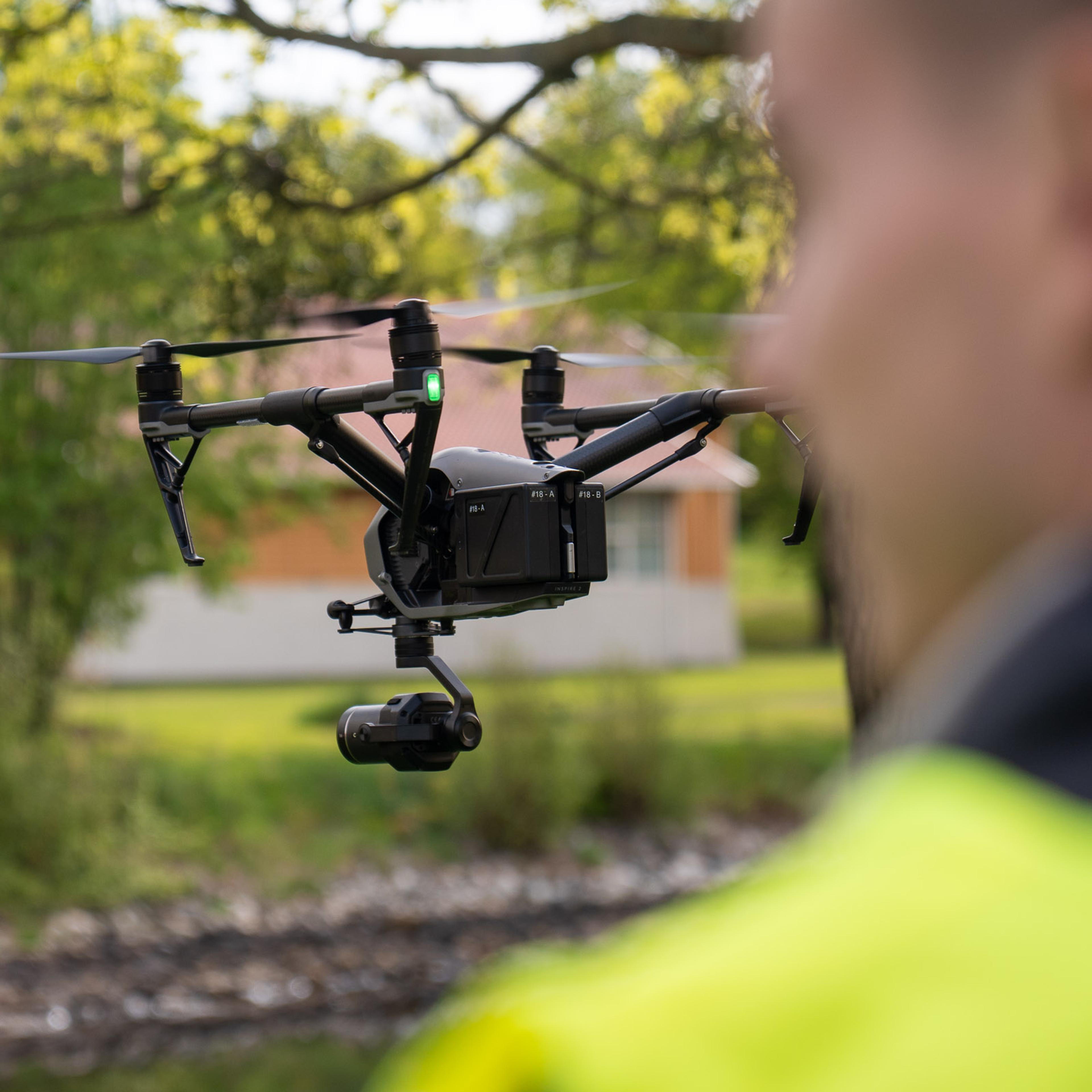 Drone-operatør i gul refleksvest styrer en svart multikopter over grønt landskap med bygning i bakgrunnen.