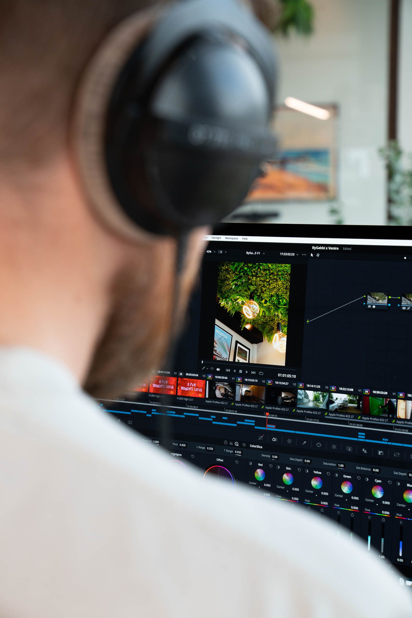 Videoproduktør med hodetelefoner jobber ved skjerm med videoedigeringsprogram og farvekorrigeringspanel.