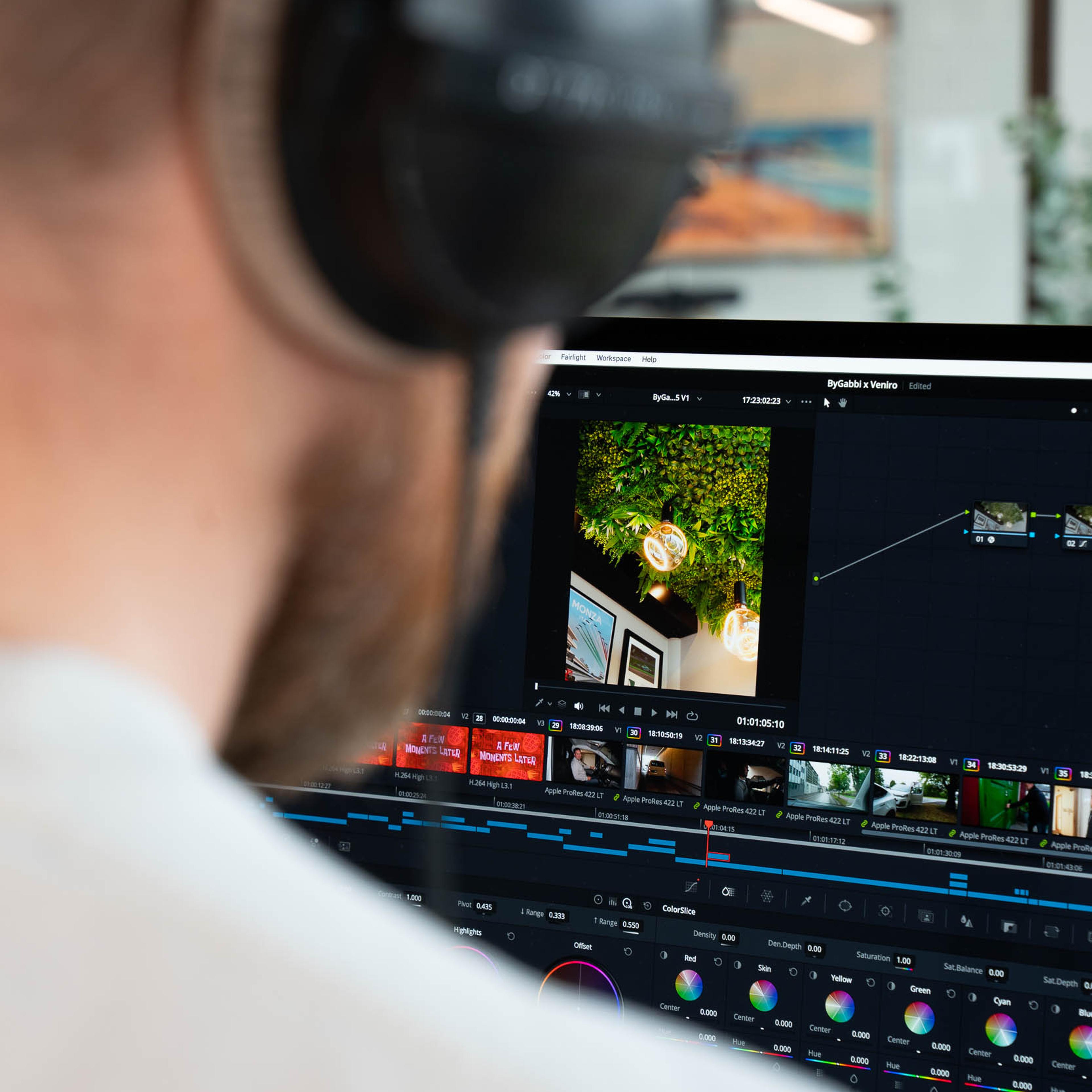 Videoproduktør med hodetelefoner jobber ved skjerm med videoedigeringsprogram og farvekorrigeringspanel.