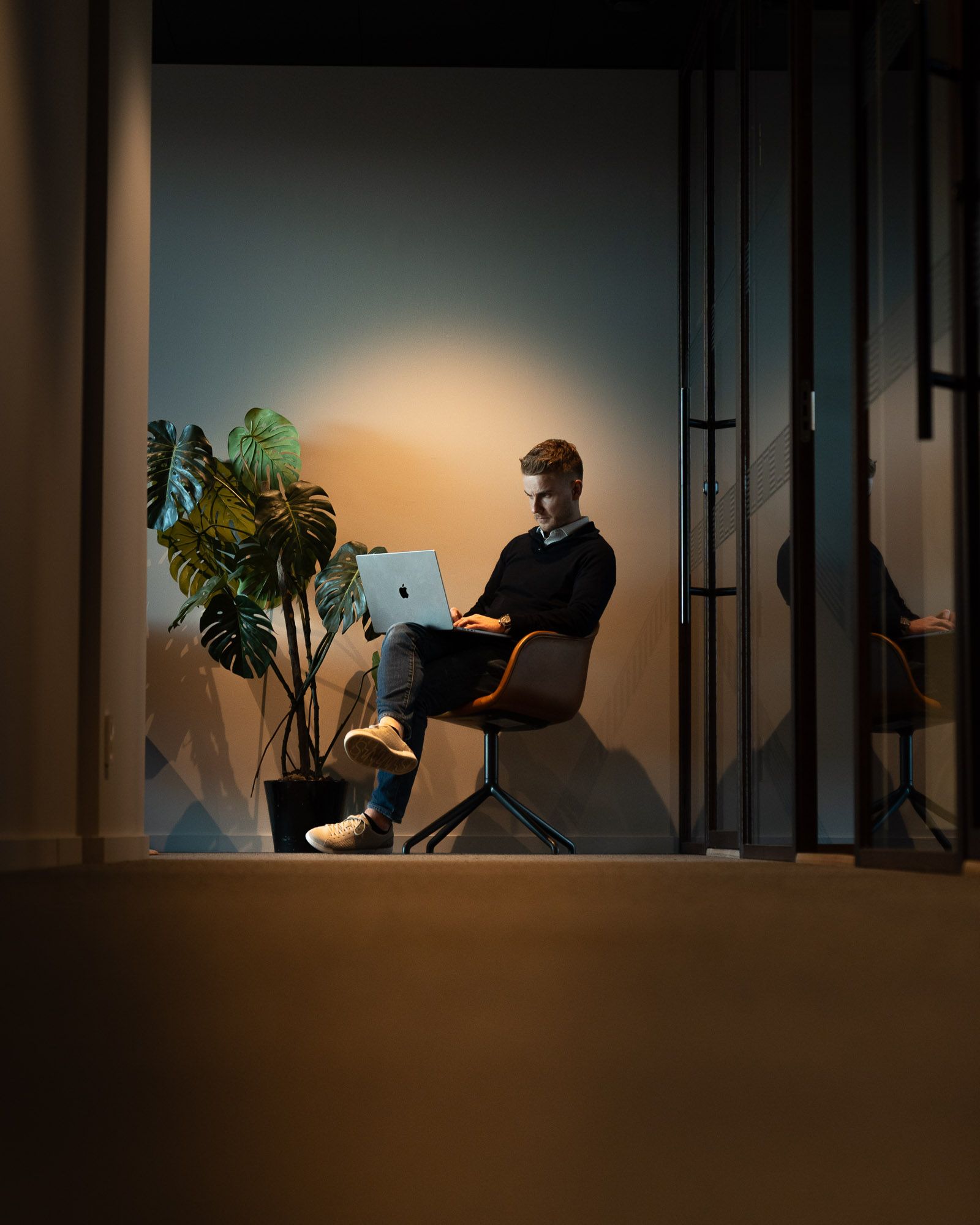 Mann arbeider på MacBook i minimalistisk kontor med plante og varmt lys, sittende i orange stol ved glassdør.
