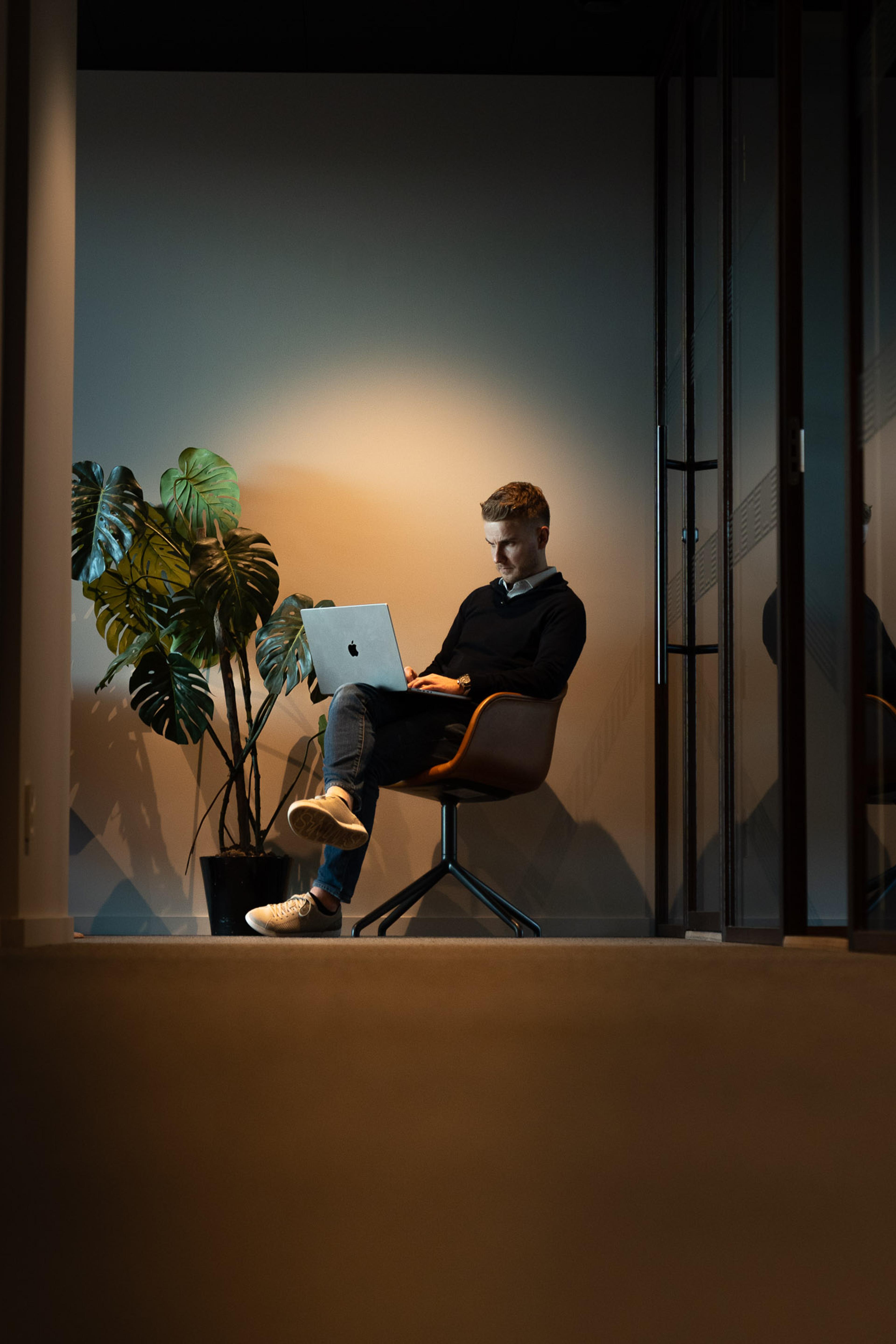 Mann arbeider på MacBook i minimalistisk kontor med plante og varmt lys, sittende i orange stol ved glassdør.