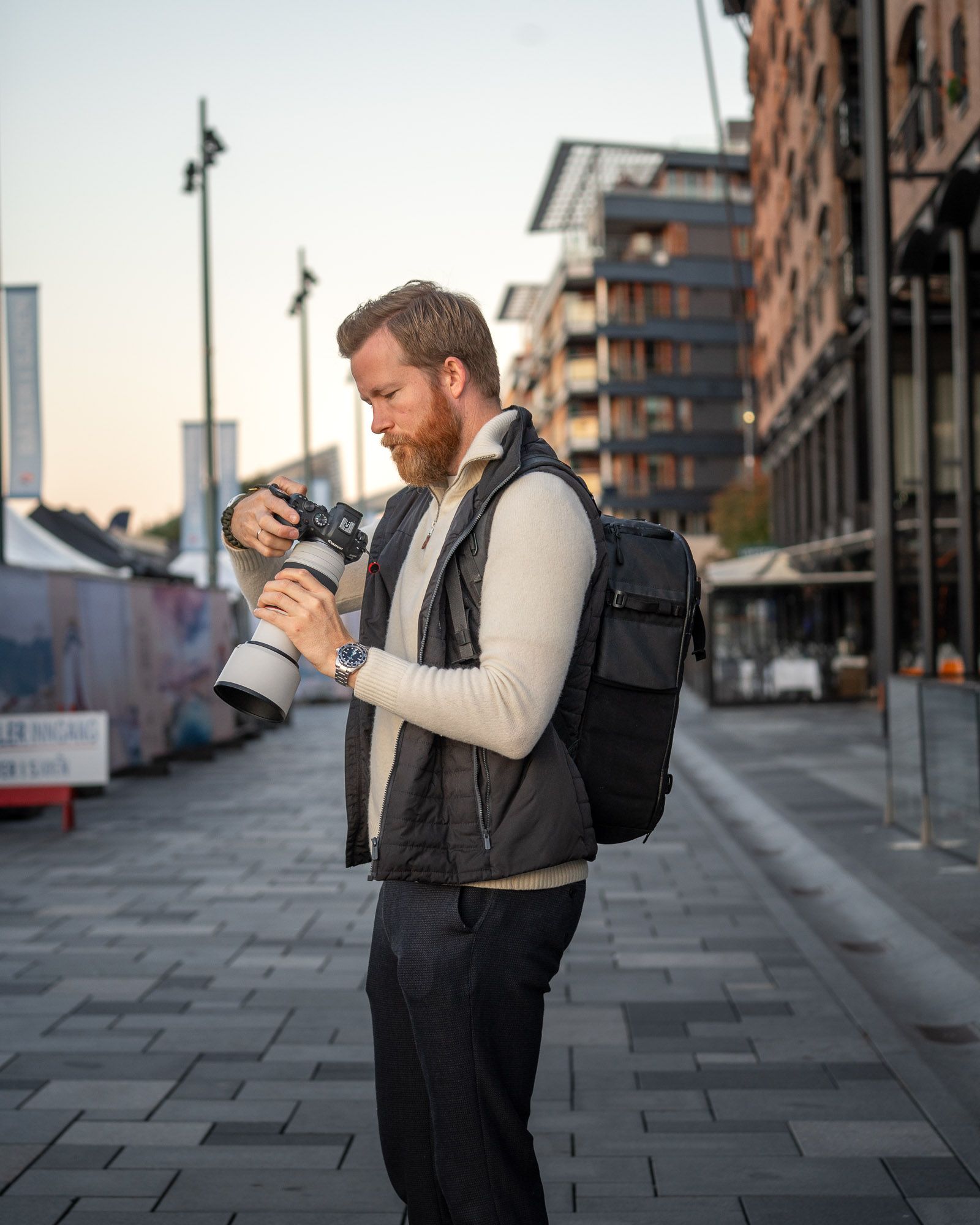 Mann med kamera og teleobjektiv på urbant torg omgitt av moderne bygninger, klar til å fotografere.