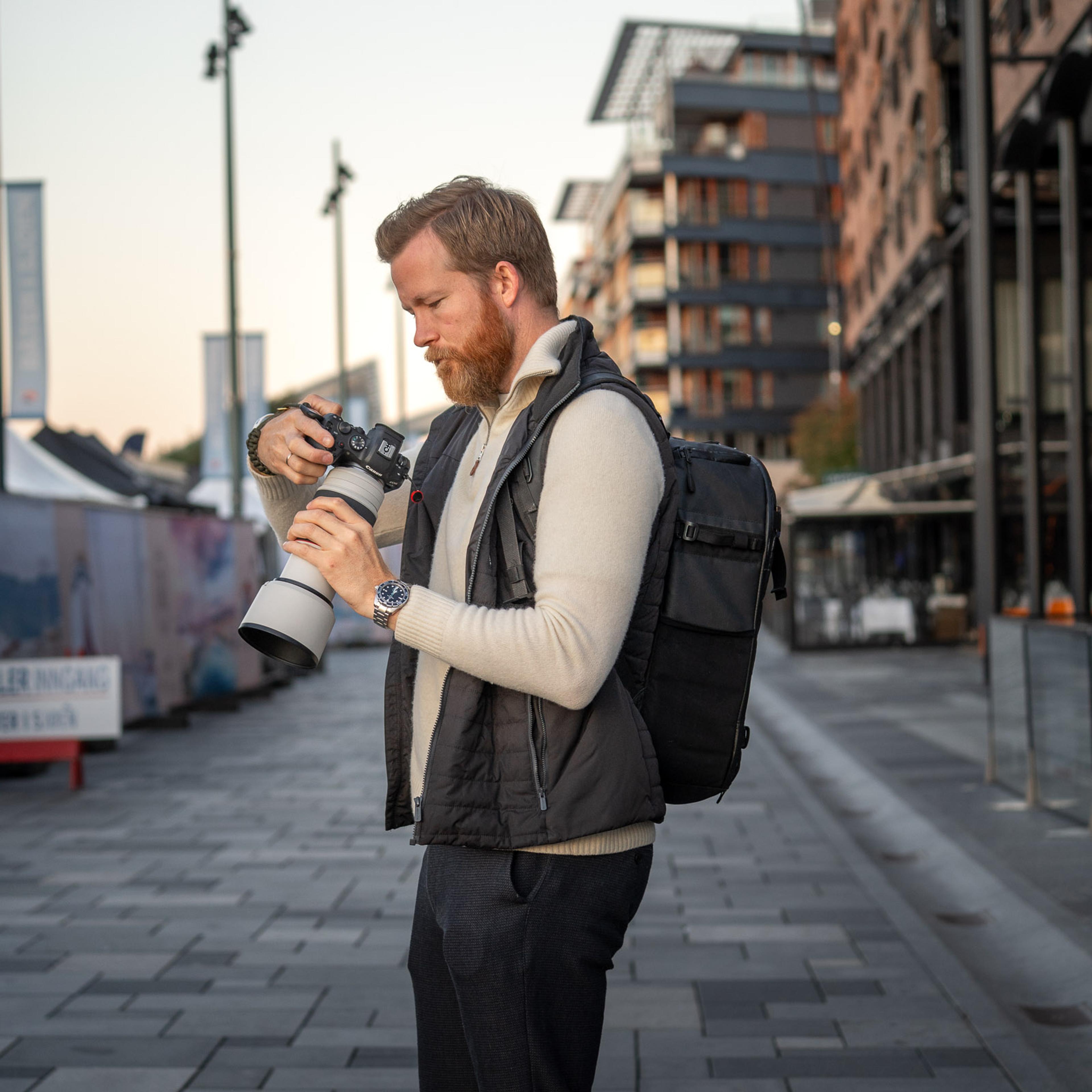 Mann med kamera og teleobjektiv på urbant torg omgitt av moderne bygninger, klar til å fotografere.