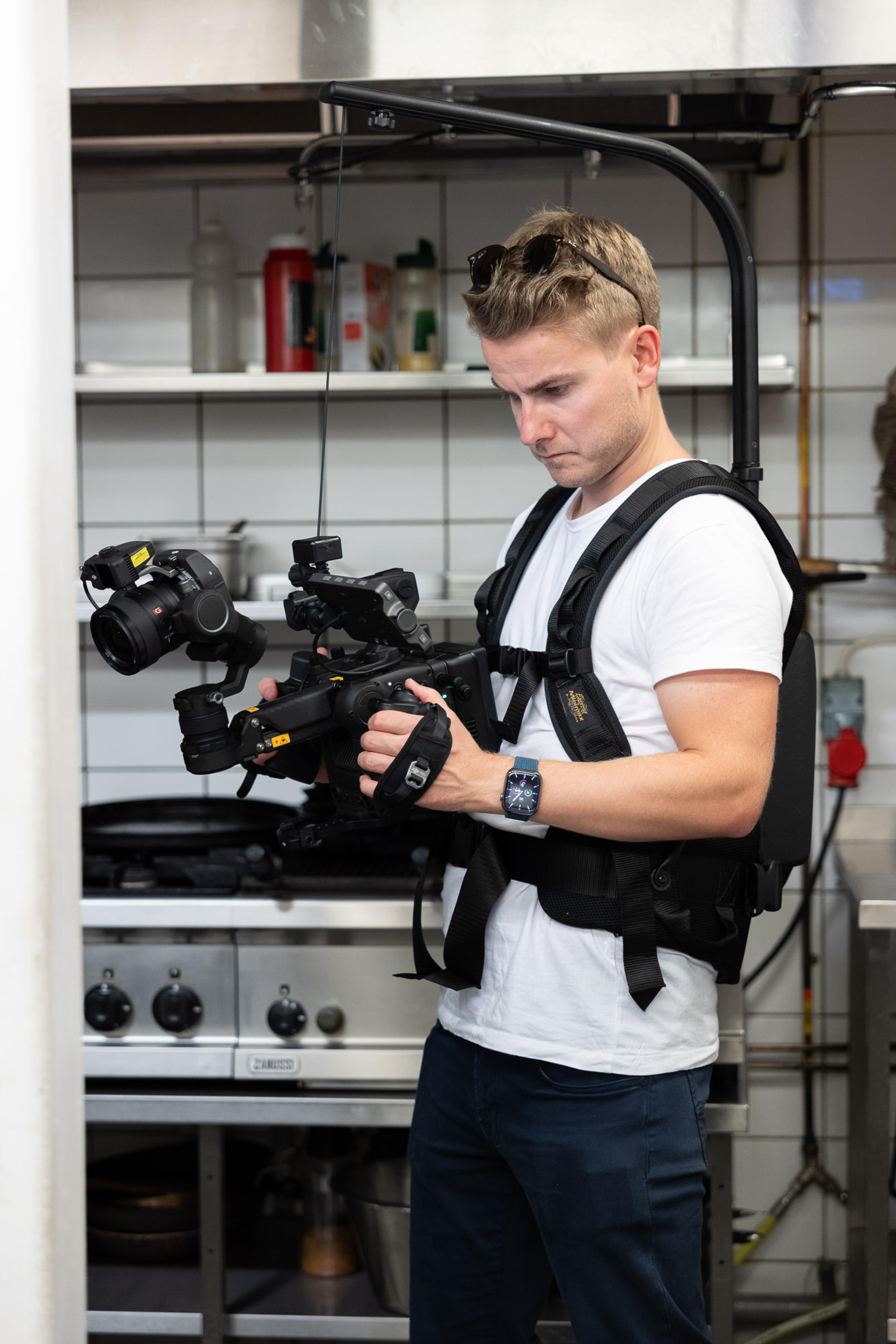 Videograf holder kamera på gimbal-stabilisator i professionelt oppsett med studio-belysning i bakgrunnen.