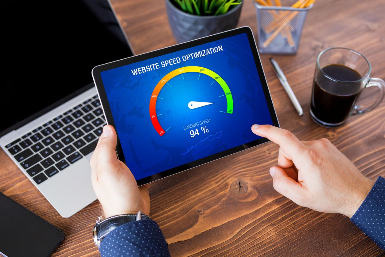 Person holder tablet som viser «Website Speed Optimization» med hastighetsmåler på 94 prosent, ved skrivebord med laptop og kaffe.