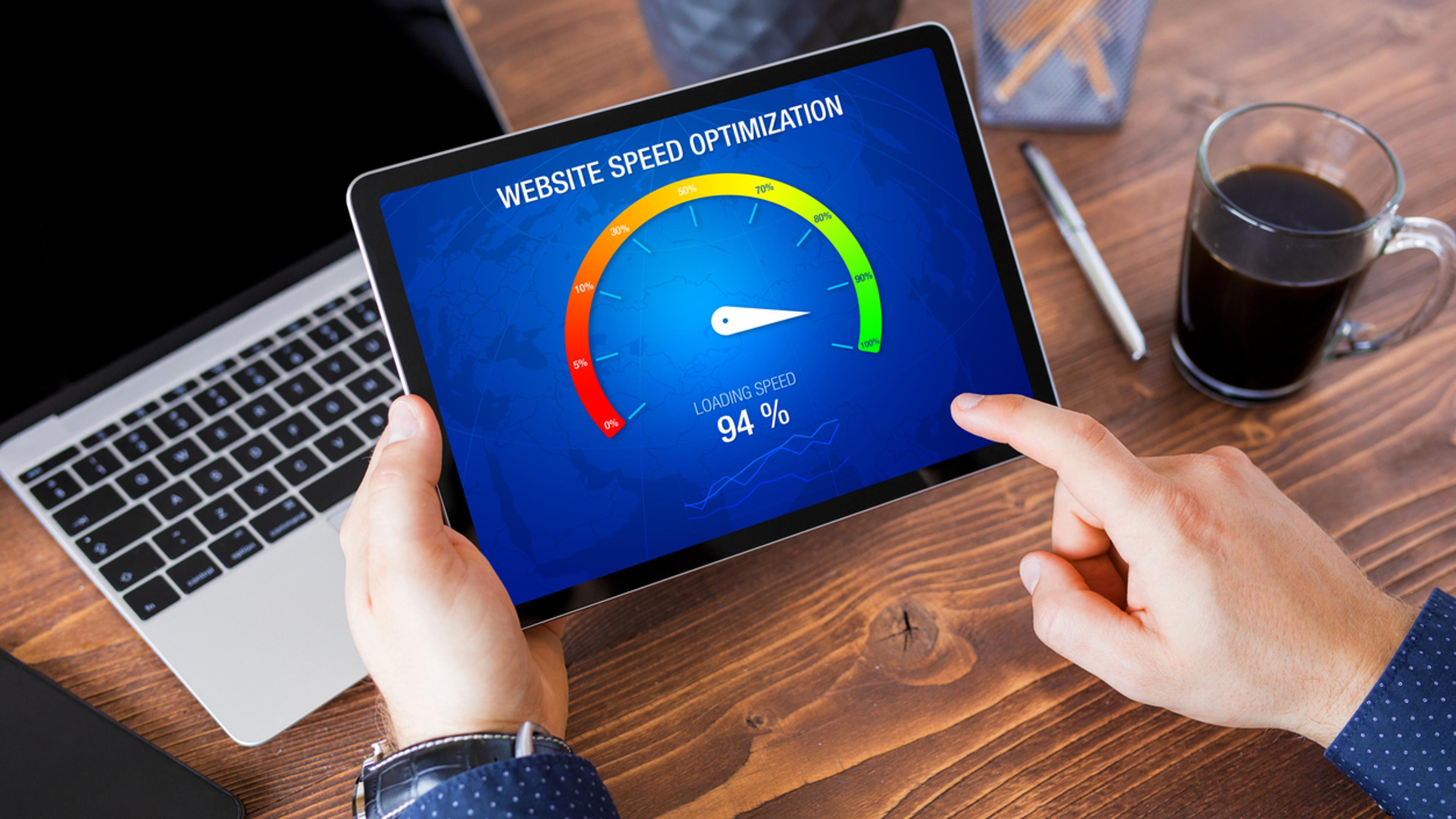 Person holder tablet som viser «Website Speed Optimization» med hastighetsmåler på 94 prosent, ved skrivebord med laptop og kaffe.