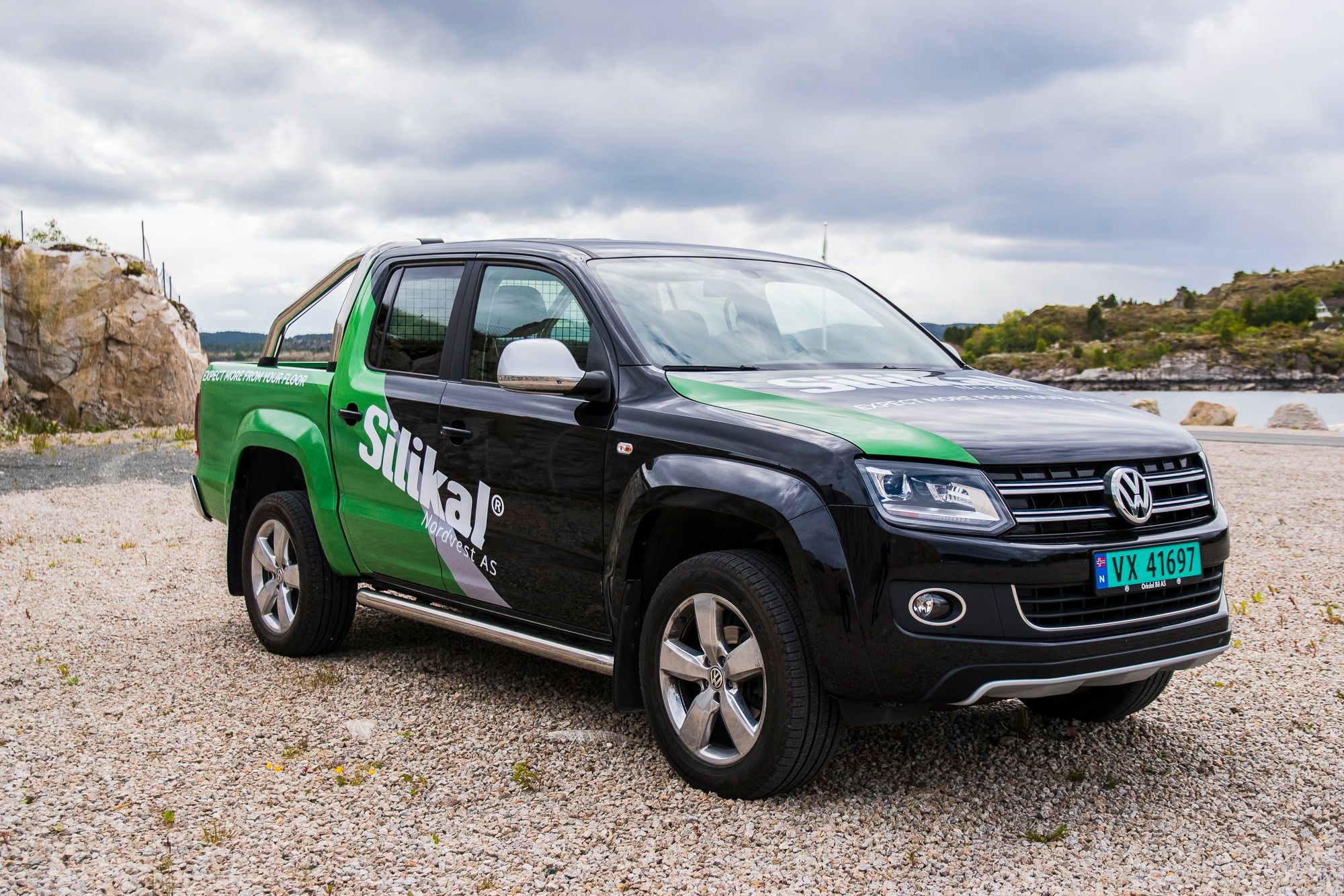 Volkswagen Amarok pickup truck med grønn og svart Sirikal-folie parkert på grusområde ved strand.