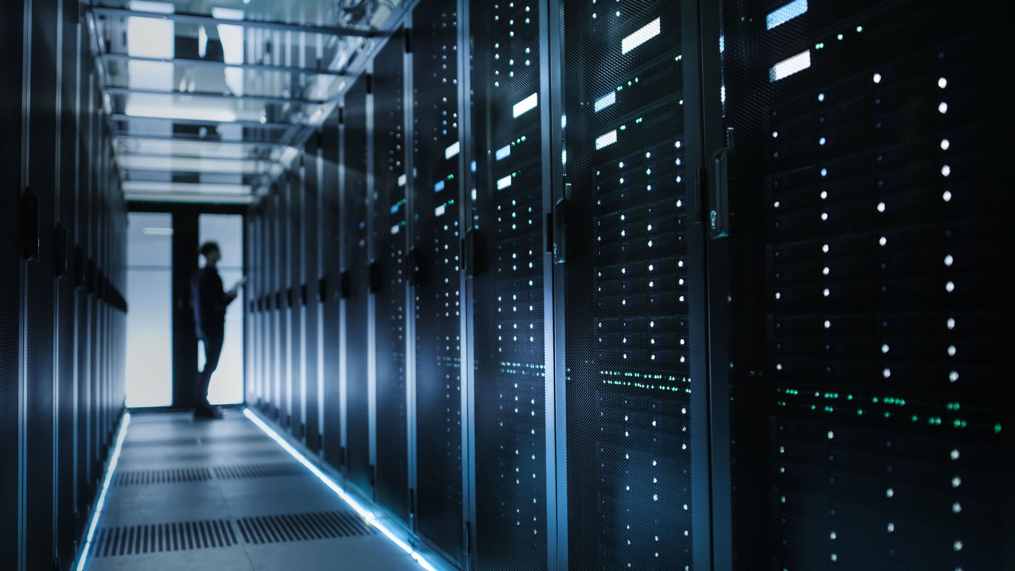Tekniker inspiserer serverutstyr i en moderne serverhall med belyst gulv og lysende indikatorer på serverrack.