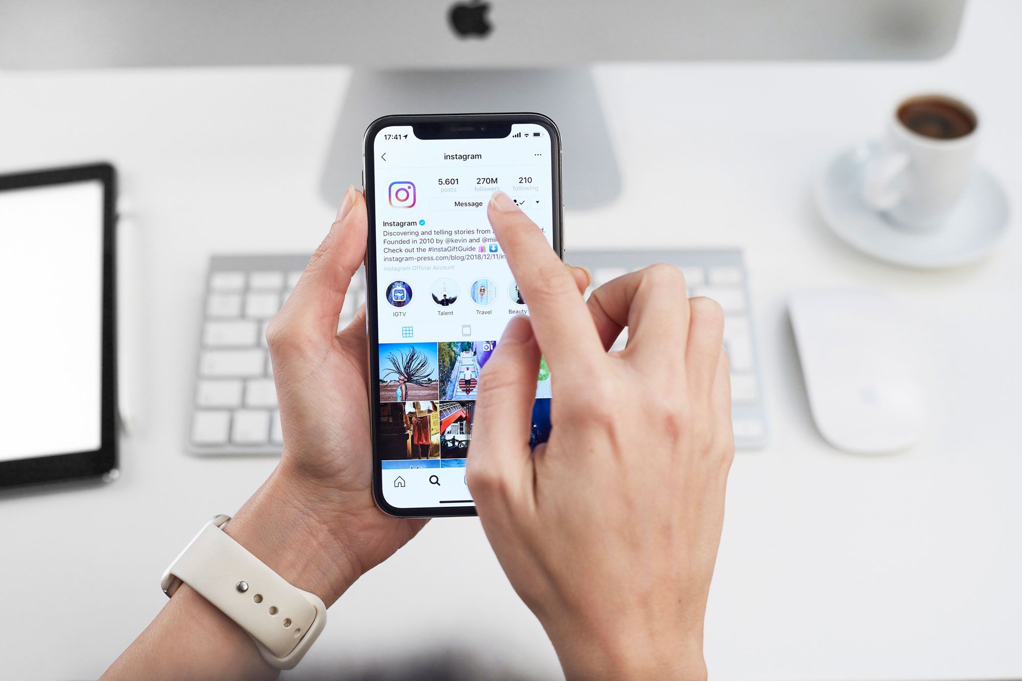 Person holder smarttelefon som viser Instagram-profil med følgere, meldinger og fotogalleri på skrivebord med dator og kaffe.