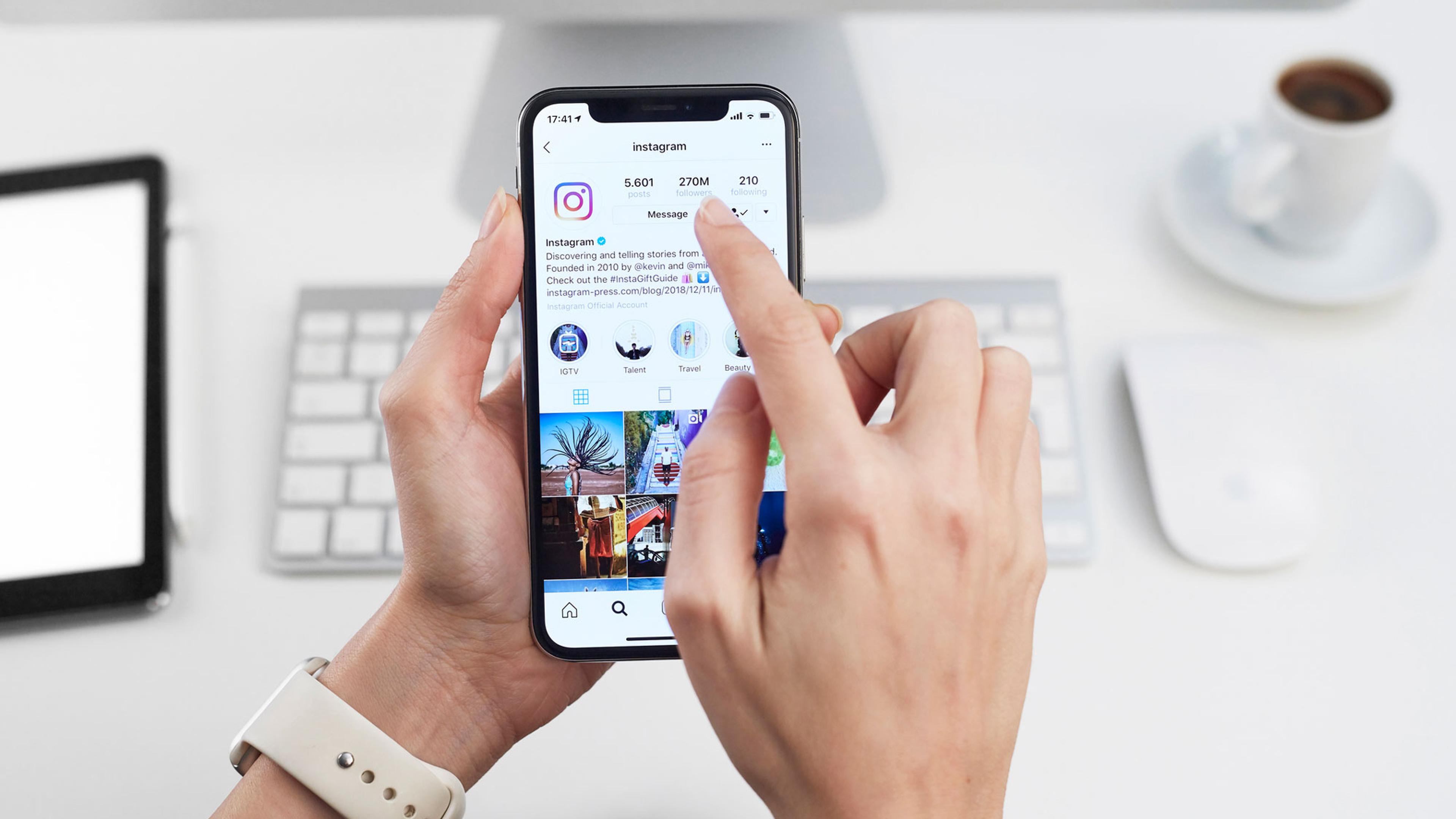 Person holder smarttelefon som viser Instagram-profil med følgere, meldinger og fotogalleri på skrivebord med dator og kaffe.