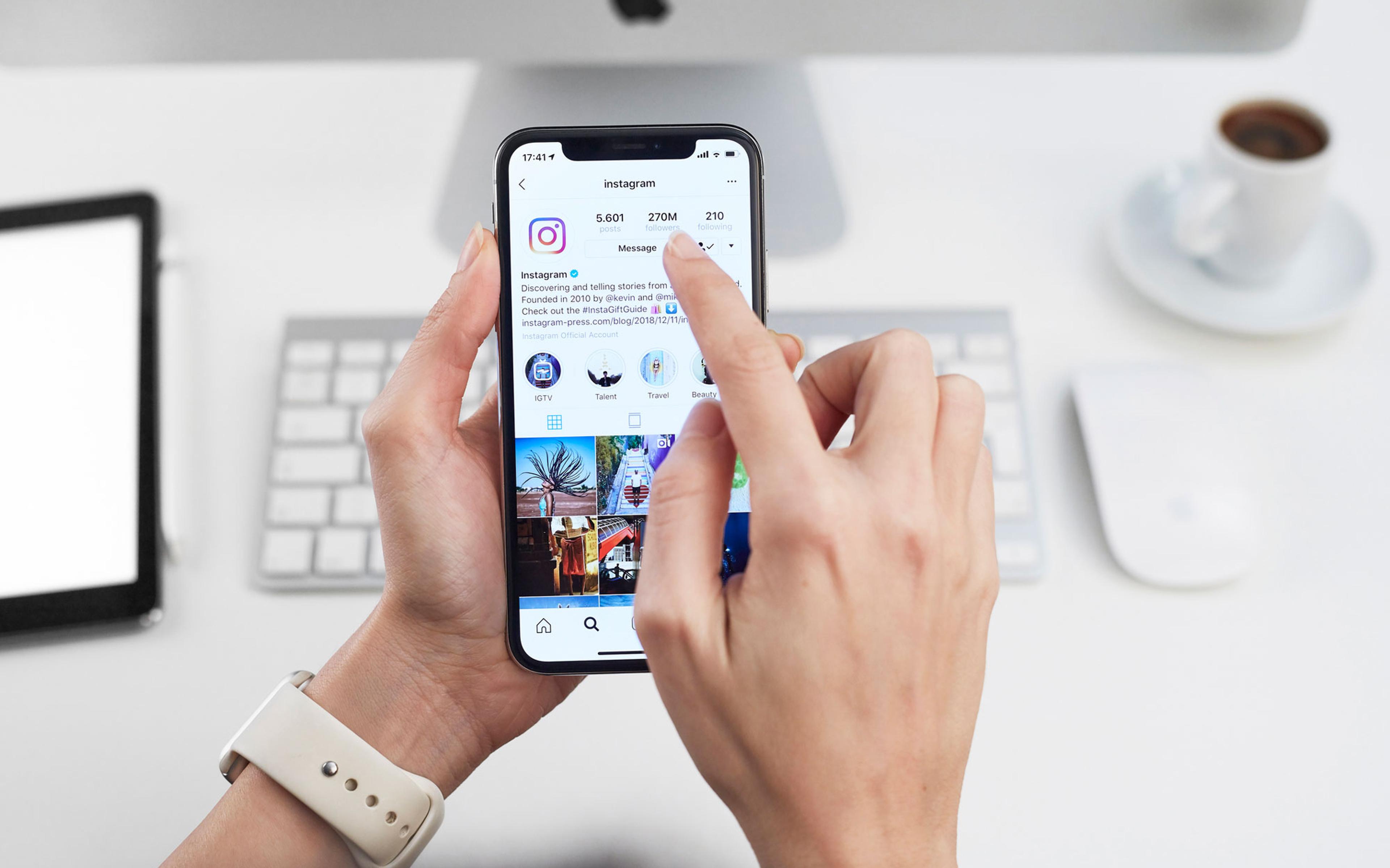 Person holder smarttelefon som viser Instagram-profil med følgere, meldinger og fotogalleri på skrivebord med dator og kaffe.