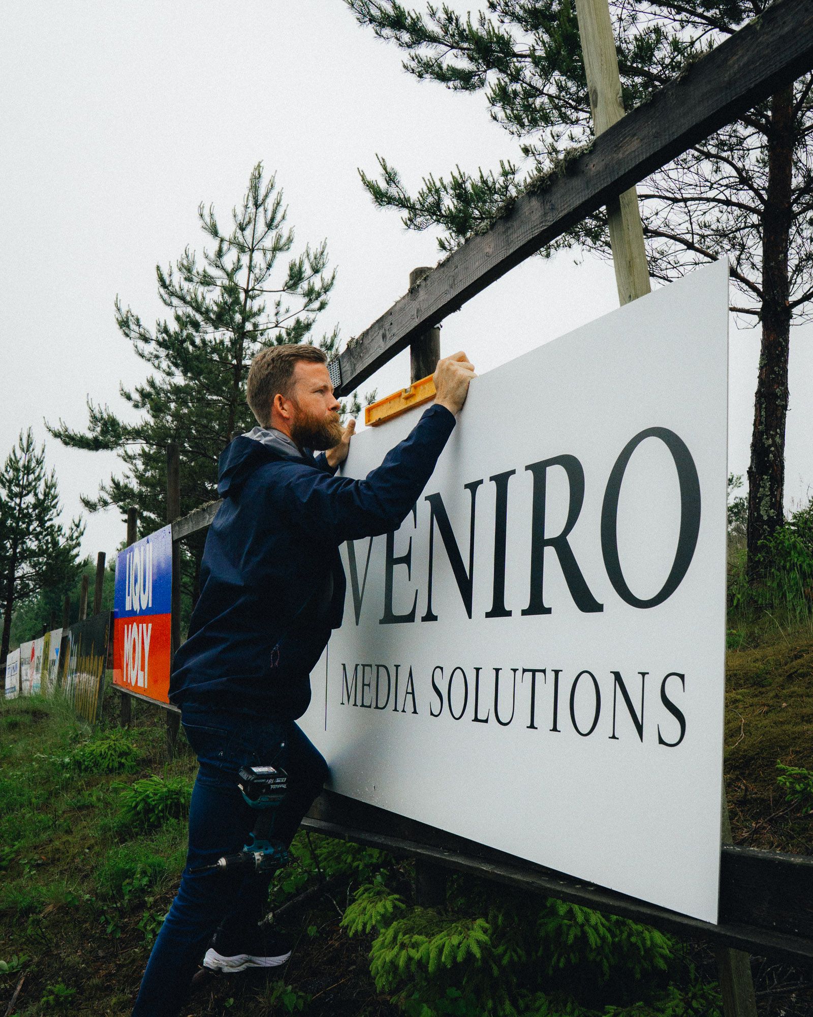 Mann monterer Veniro Media Solutions skilt på trepanel ved siden av fyrretræer i uteområde.