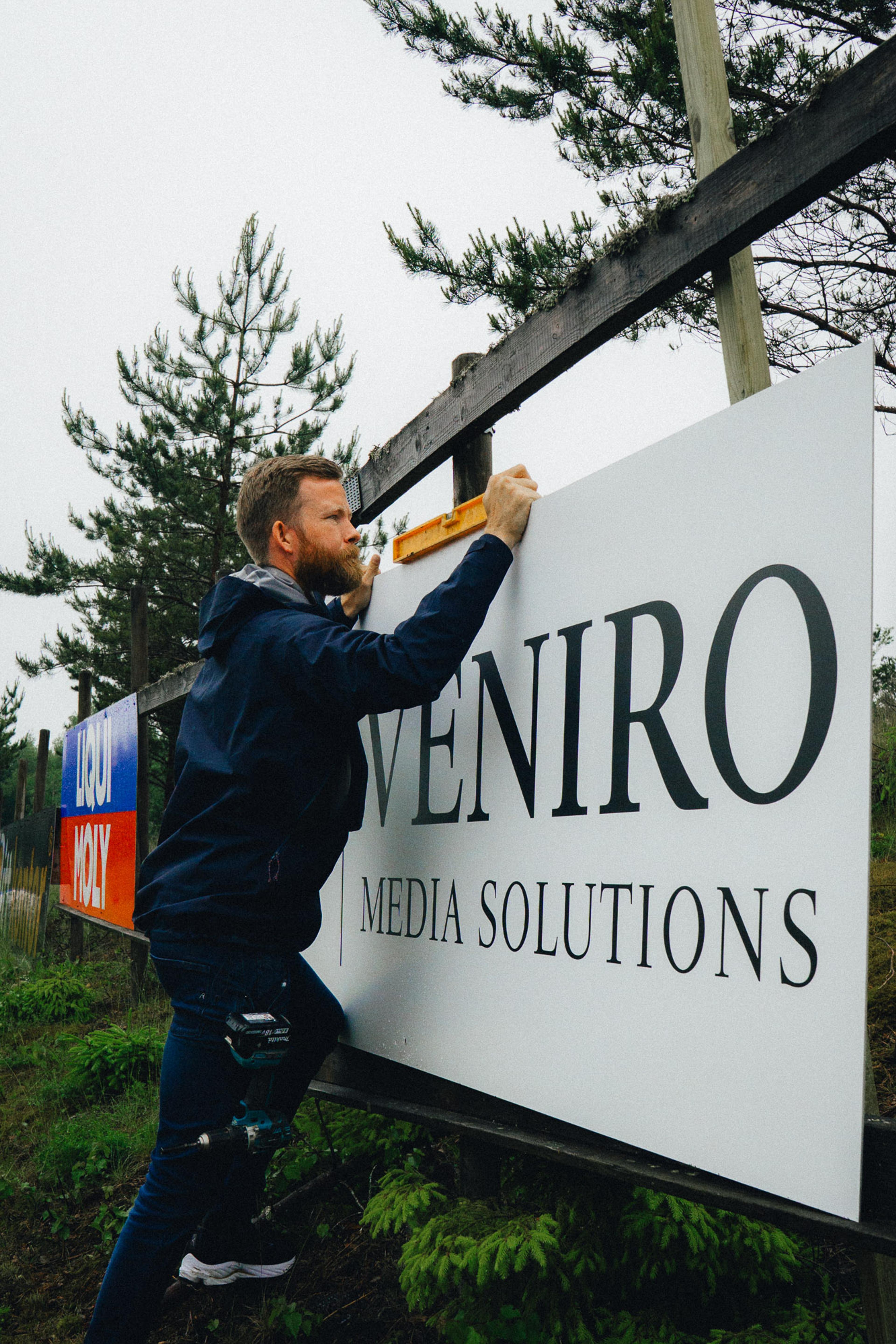 Mann monterer Veniro Media Solutions skilt på trepanel ved siden av fyrretræer i uteområde.