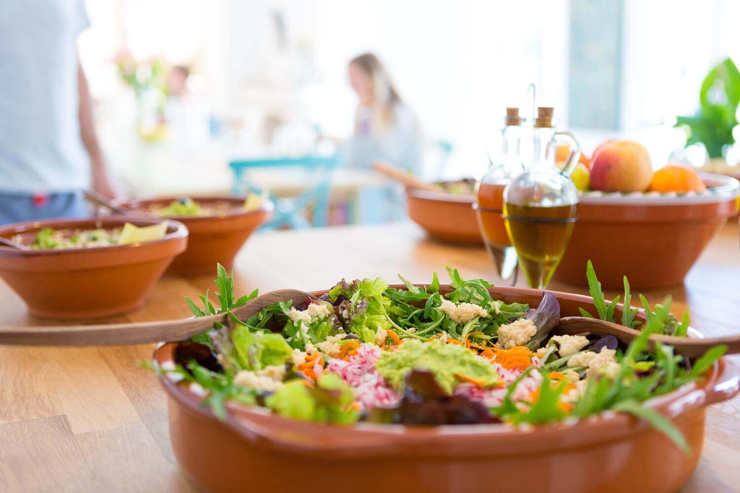 Fargerik salat i terrakottaskål på middagsbord med frukt, olje og mennesker i bakgrunnen.