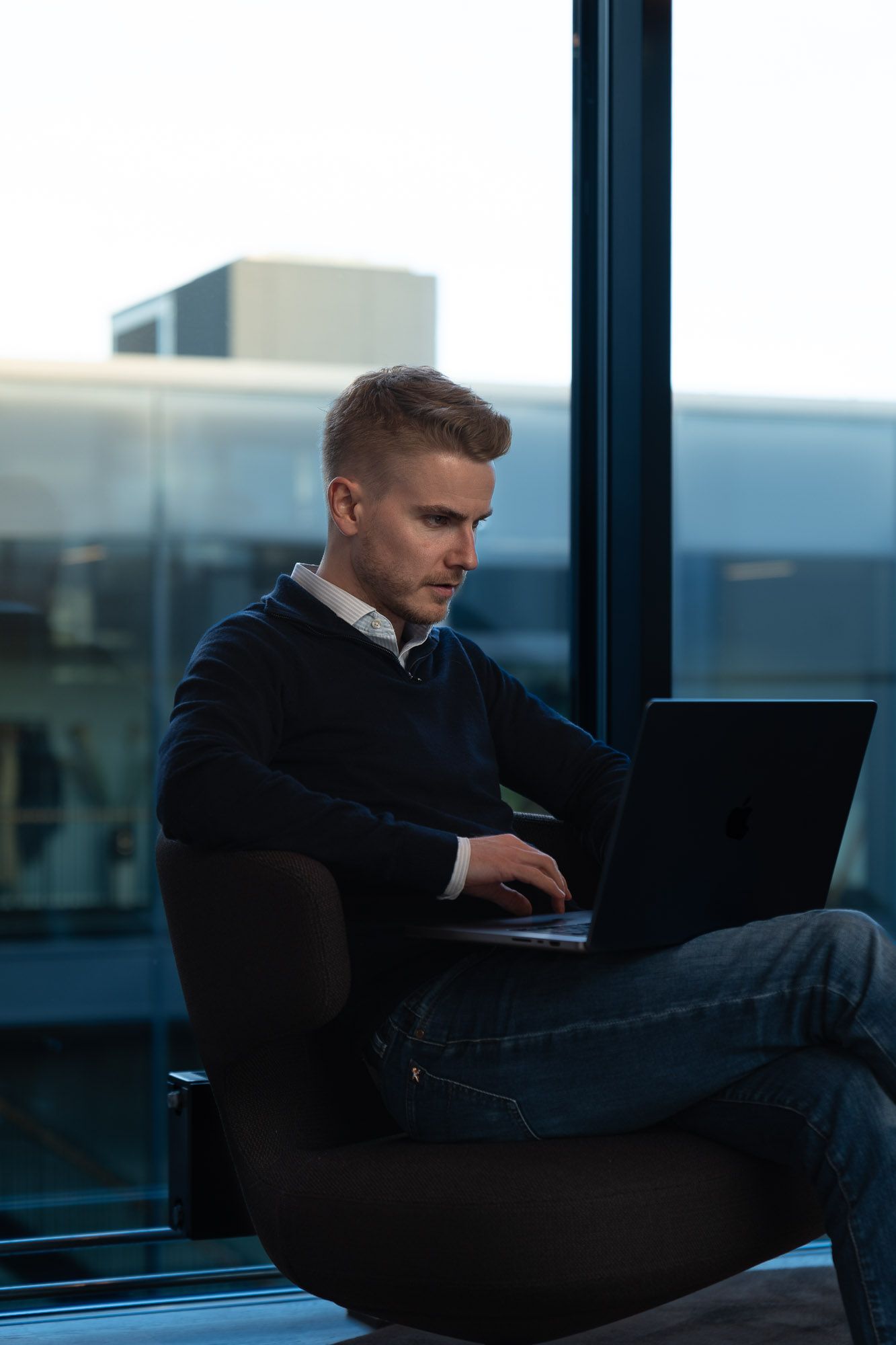 Ung mann med laptop arbeider ved stort vindu med utsikt over bylandskap og industribygg.