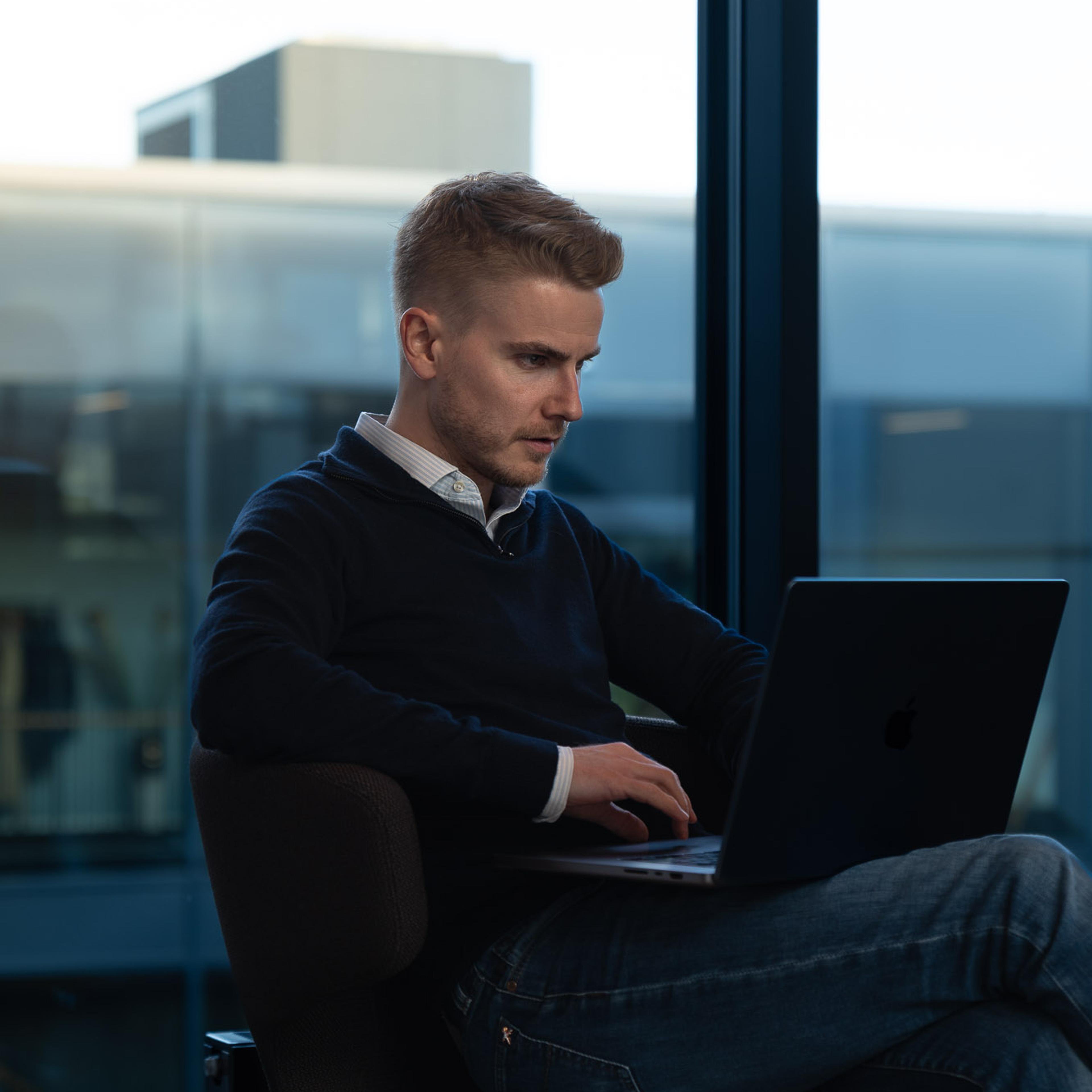 Ung mann med laptop arbeider ved stort vindu med utsikt over bylandskap og industribygg.