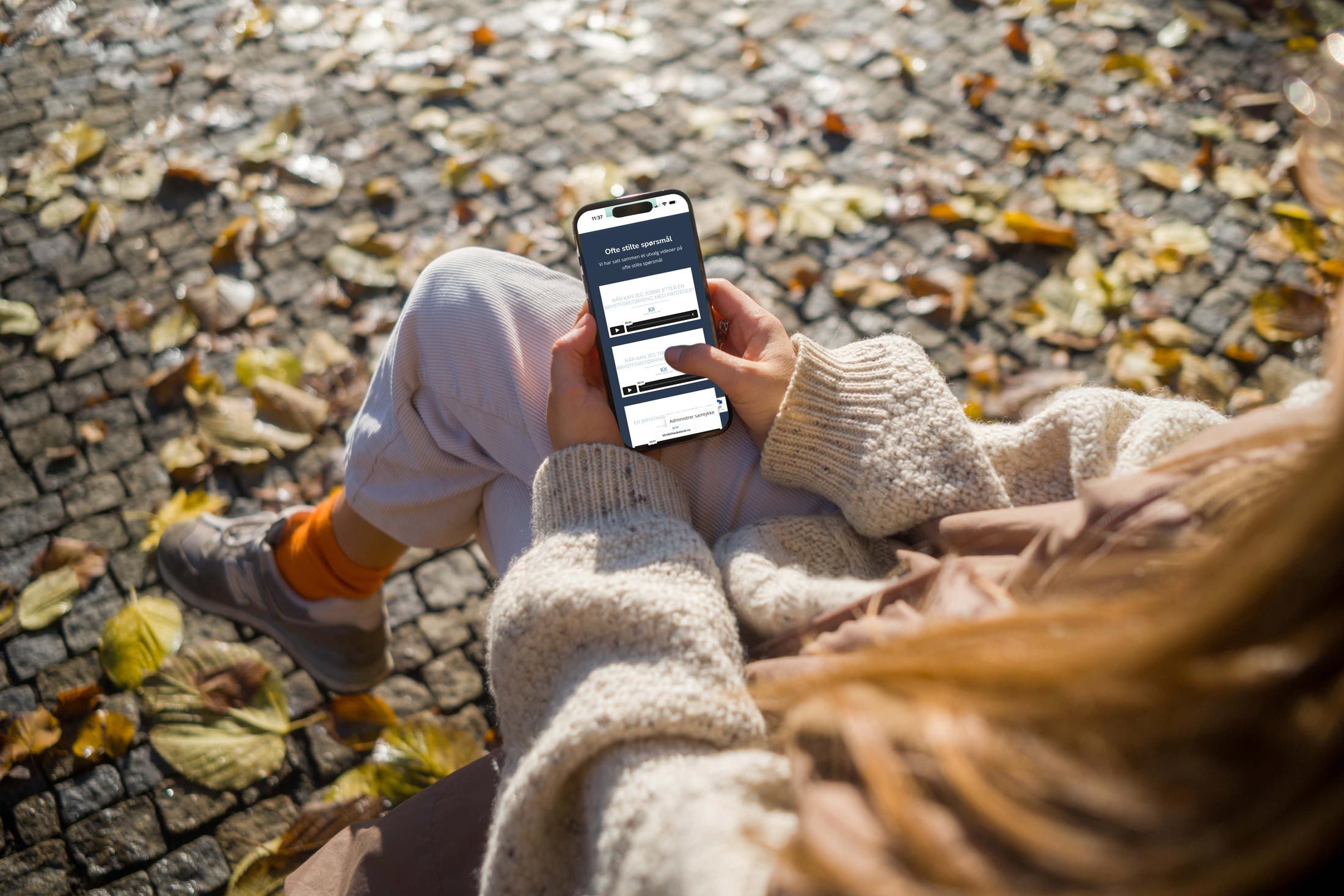 Person holder smarttelefon med applikasjonsskjerm, sitter på steinflate med høstblader rundt seg.