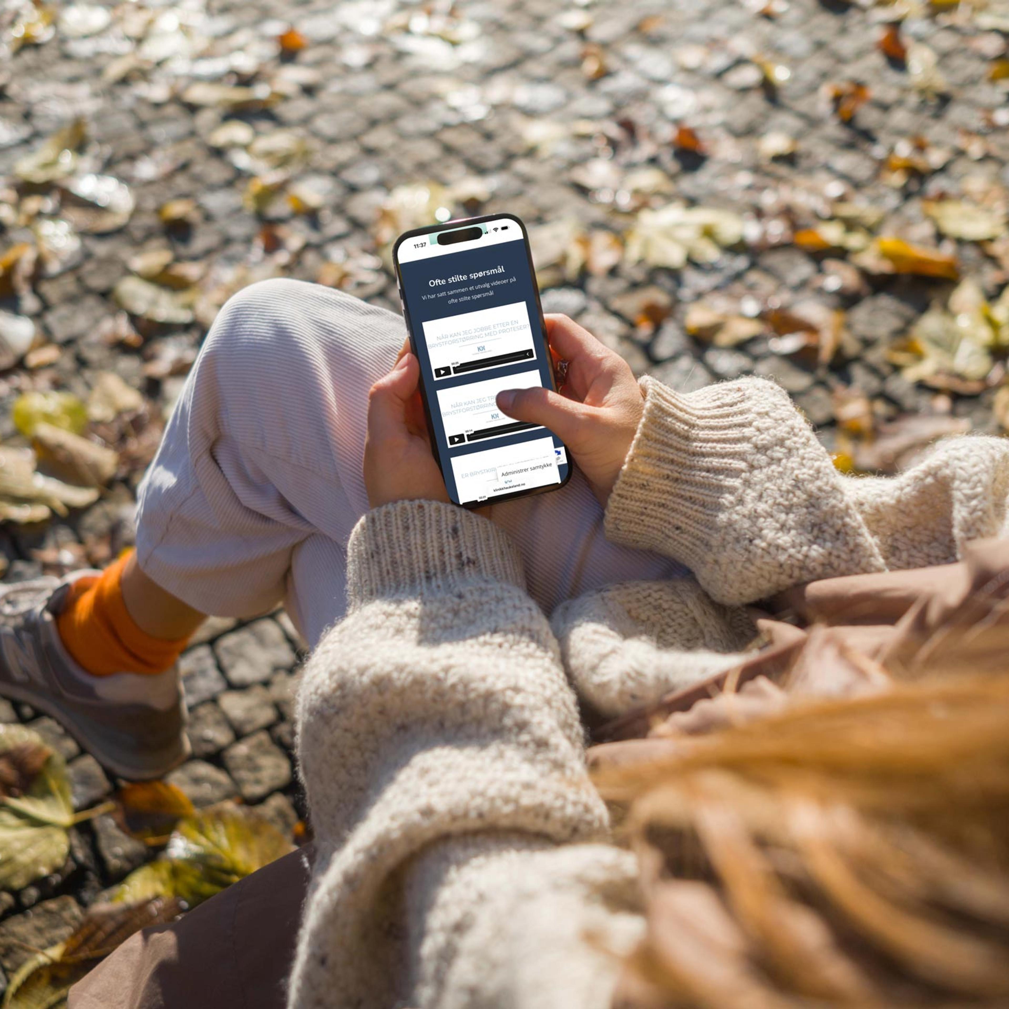 Person holder smarttelefon med applikasjonsskjerm, sitter på steinflate med høstblader rundt seg.